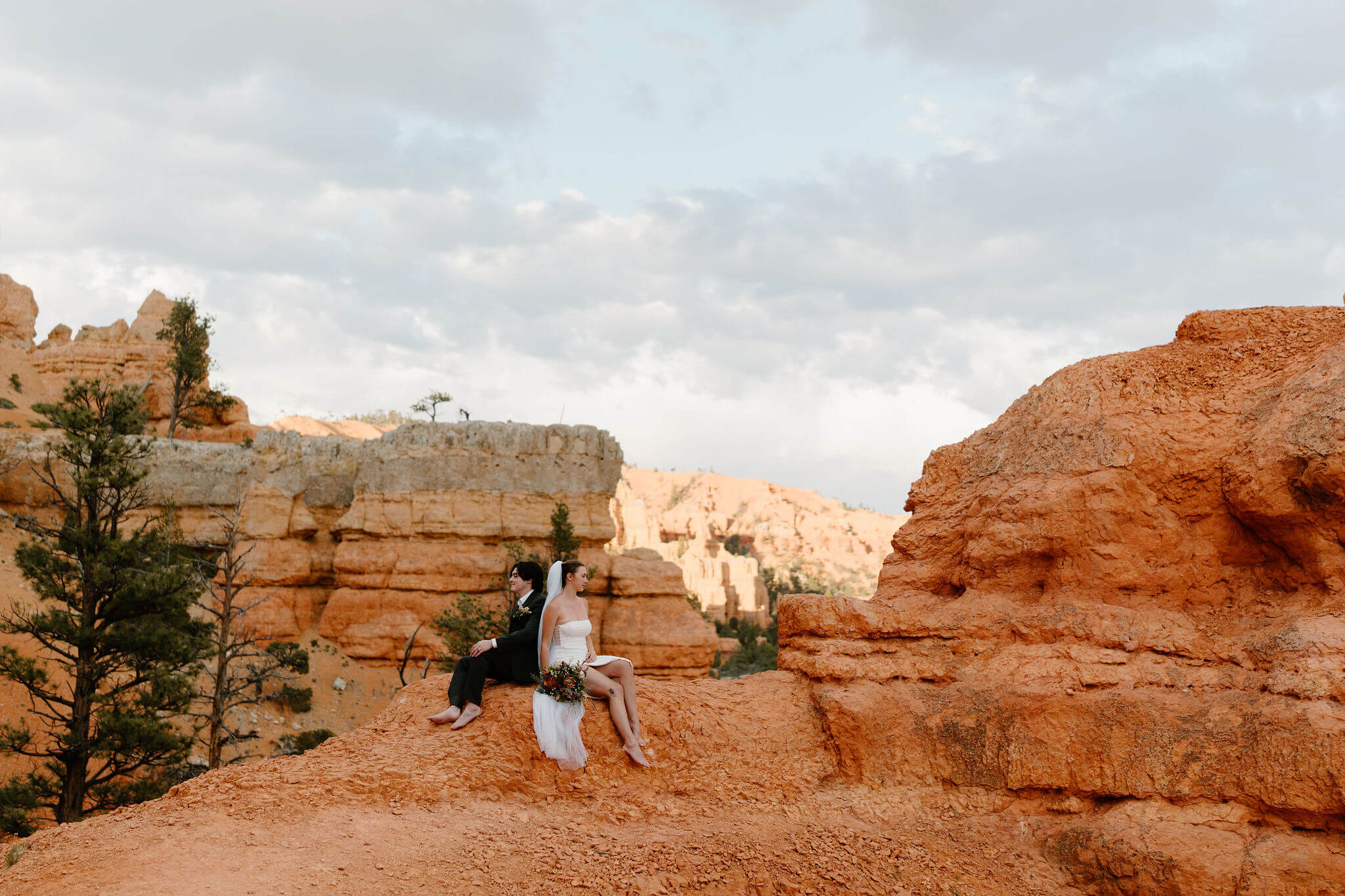 bride and groom sitting on a red rock overlook, back to back, with red cliffs all around them at the Red Canyon Trailhead