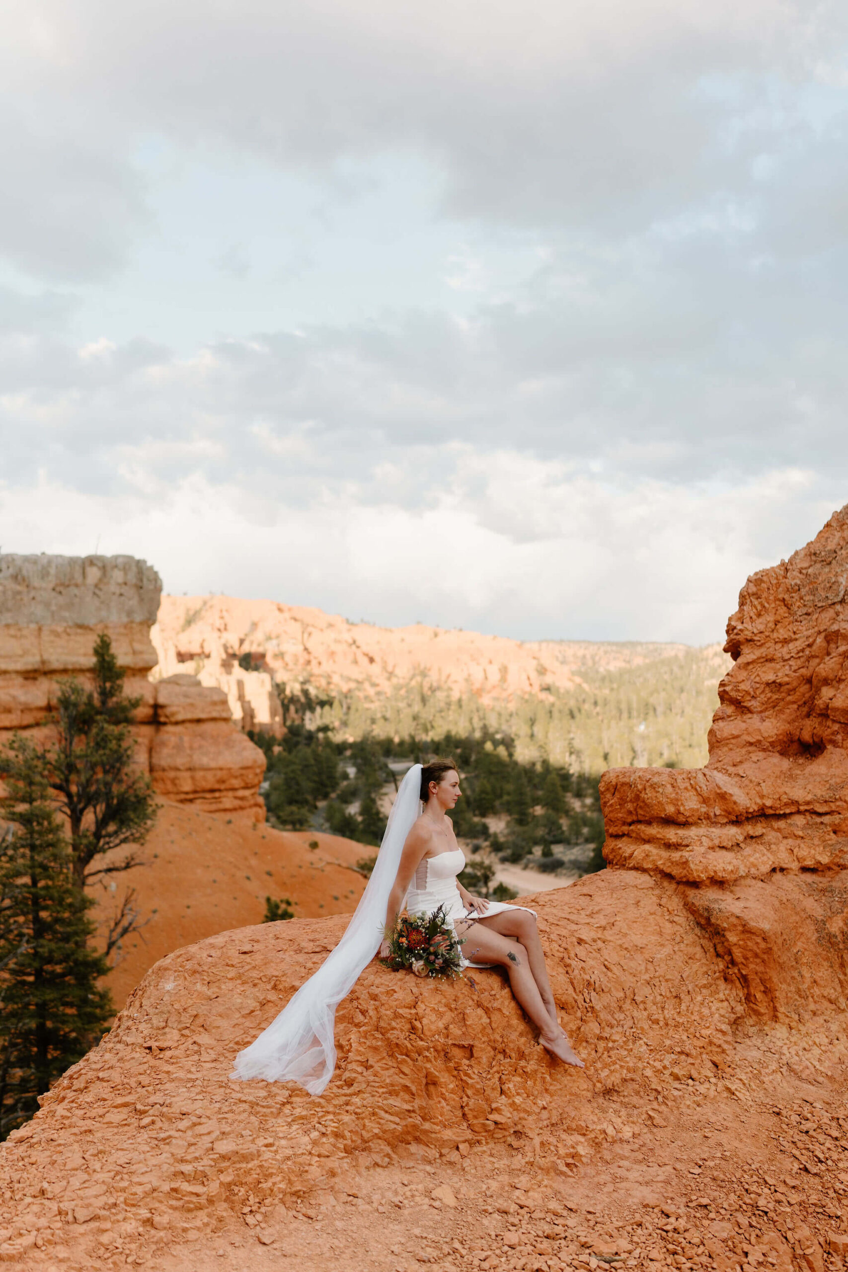 bride sitting on a cliff of red rocks in a short white dress, veil flowing behind her, looking off camera with her bouquet at her side