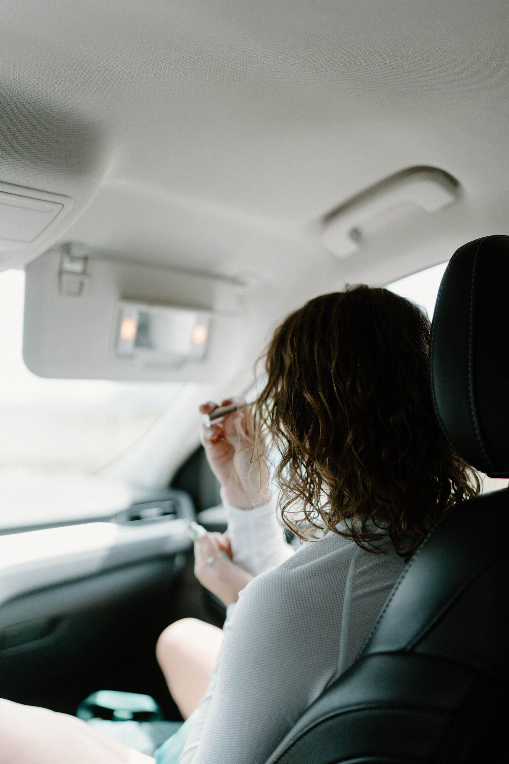 photo from the backseat of a car of a woman putting on mascara in the sun visor mirror in the passenger seat