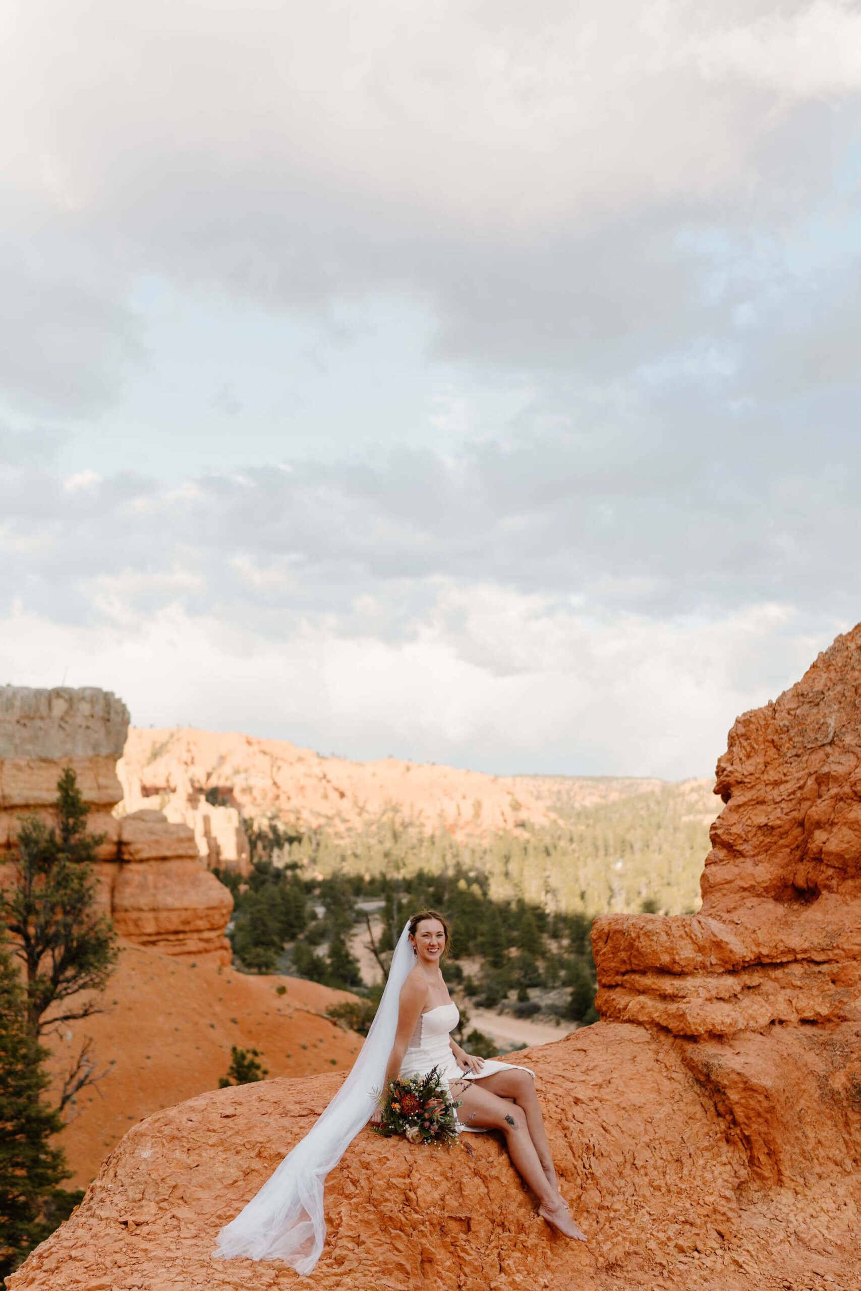 bride sitting on a cliff of red rocks in a short white dress, veil flowing behind her, smiling at the camera with her bouquet at her side