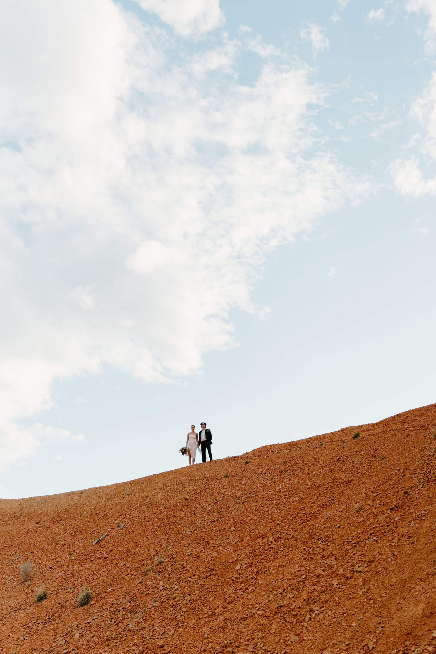 wide image of bride and groom standing shoulder-to-shoulder on top of a large hill of red rocks and stone, holding hands and looking stoically at the camera against a blue sky