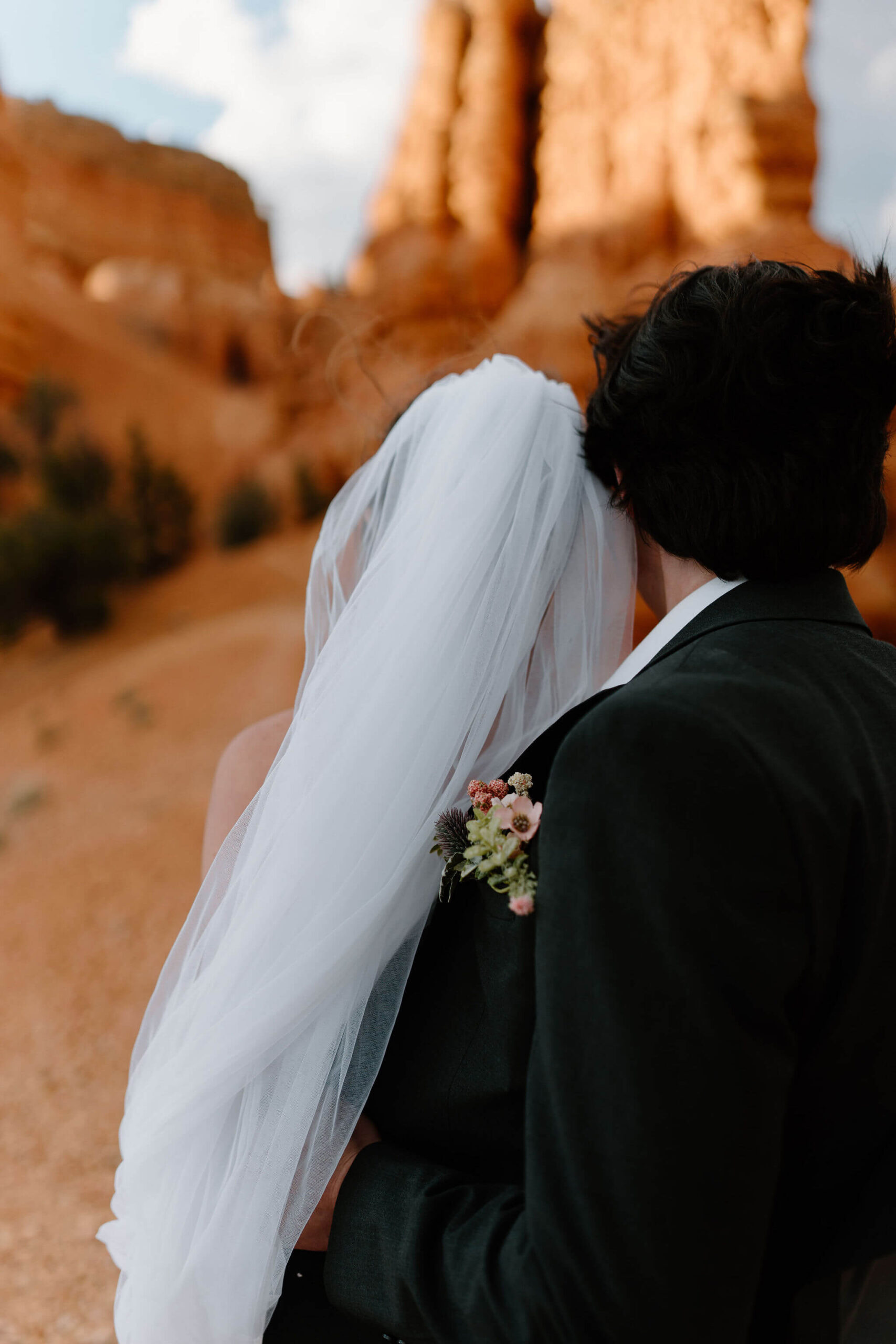 close up image of groom's boutonniere as he holds bride steady, tall red rocks in the background at the Red Canyon Trailhead