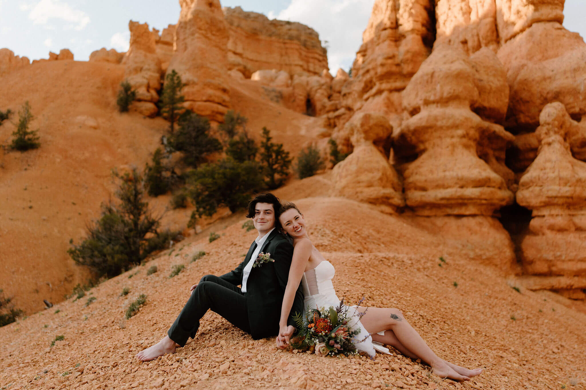 bride and groom sitting back to back at the crest of a hill on a narrow trail, red hoodoos behind them, leaning against each other and smiling toward the camera