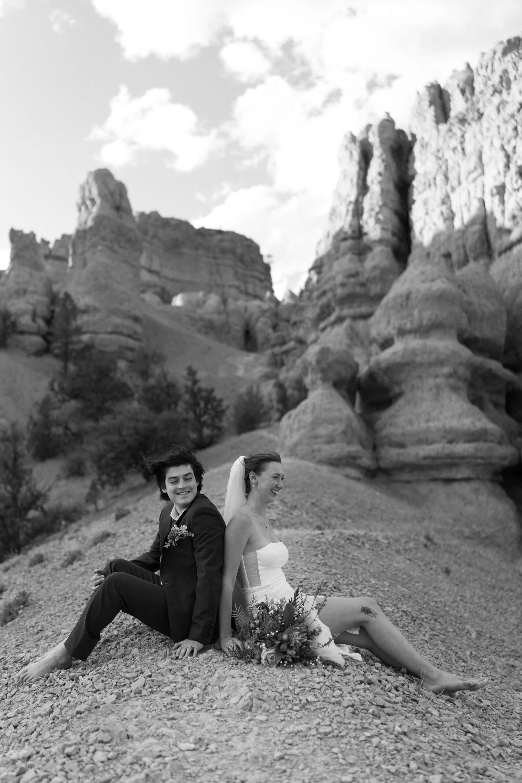 black and white image of a bride and groom sitting back to back at the crest of a hill on a narrow trail, hoodoos behind them, leaning against each other and laughing with her bridal bouquet at her side