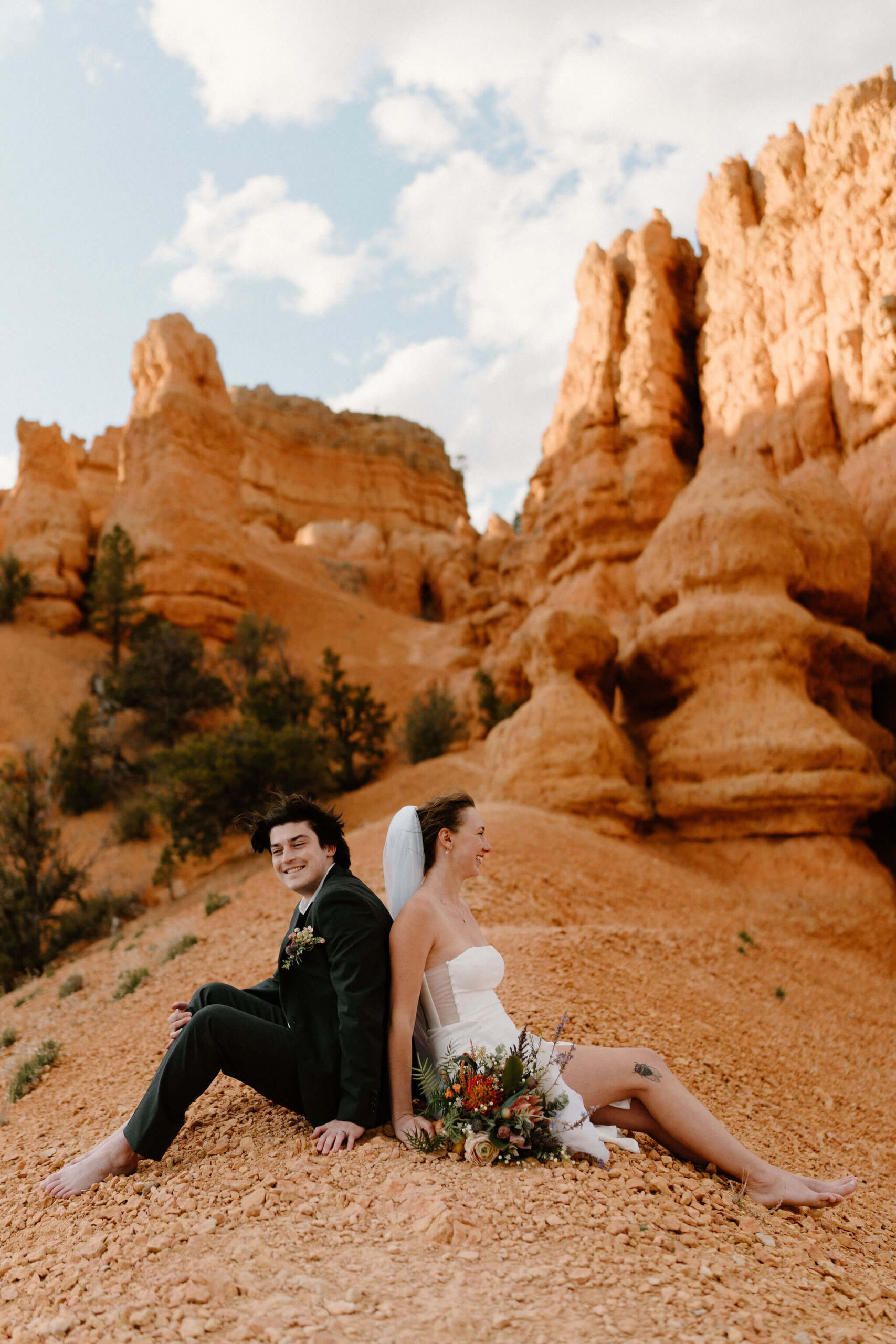 bride and groom sitting back to back at the crest of a hill on a narrow trail, red hoodoos behind them, leaning against each other and laughing with her bridal bouquet at her side