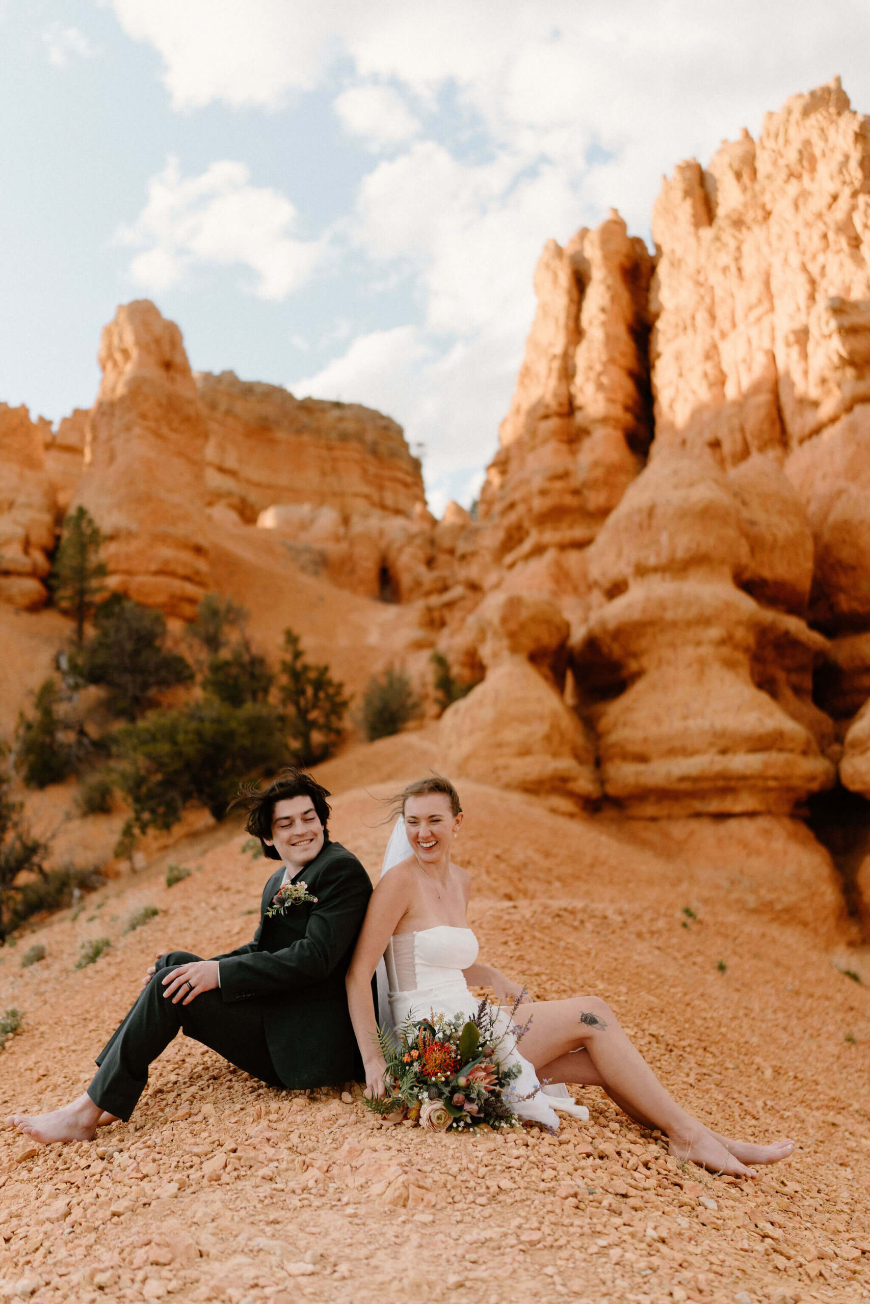 bride and groom sitting back to back at the crest of a hill on a narrow trail, red hoodoos behind them, leaning against each other and laughing with her bridal bouquet at her side