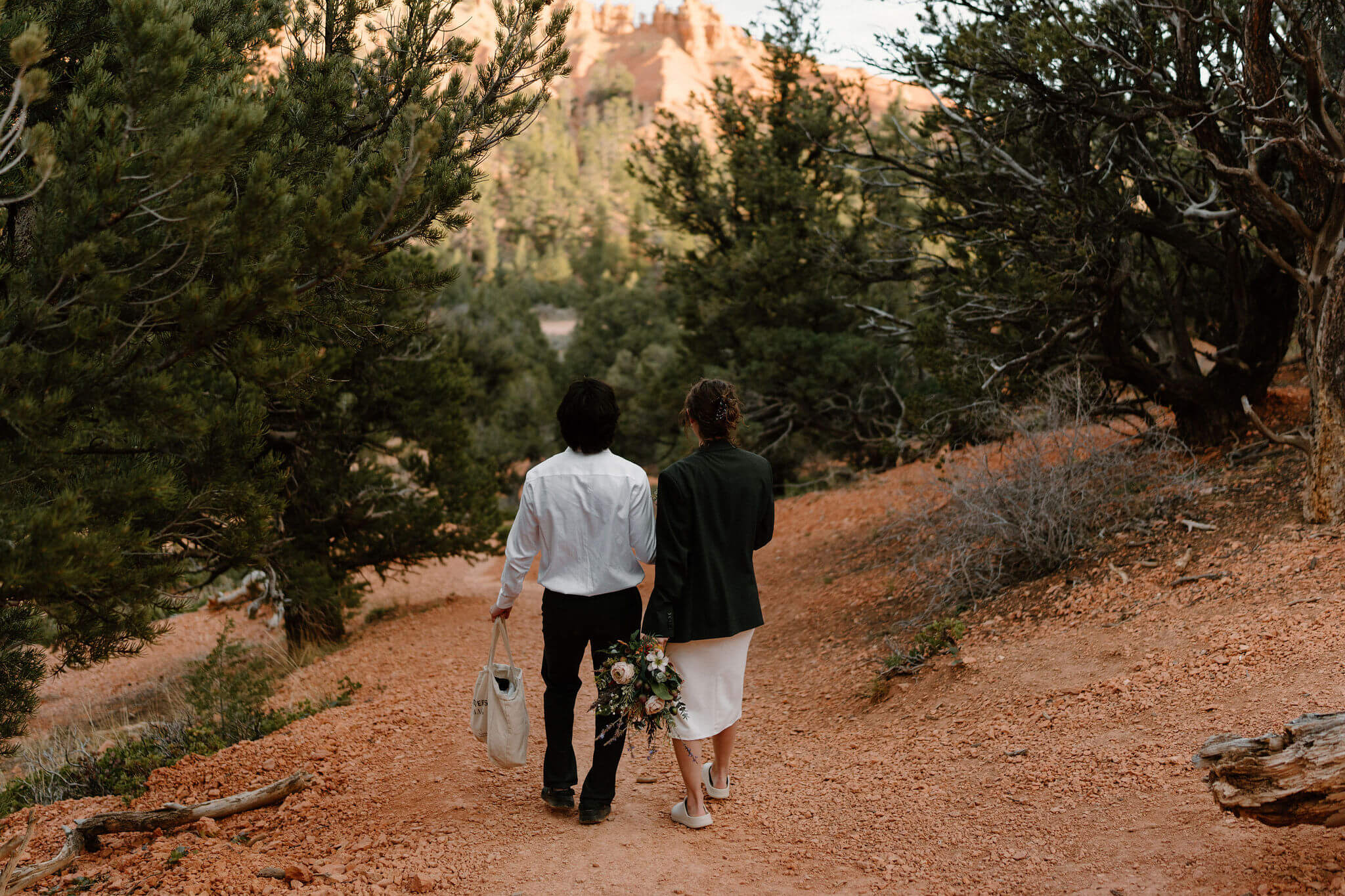 bride and groom walking down a trail, groom holding bride's tote bag and bride wearing groom's jacket and holding her bouquet