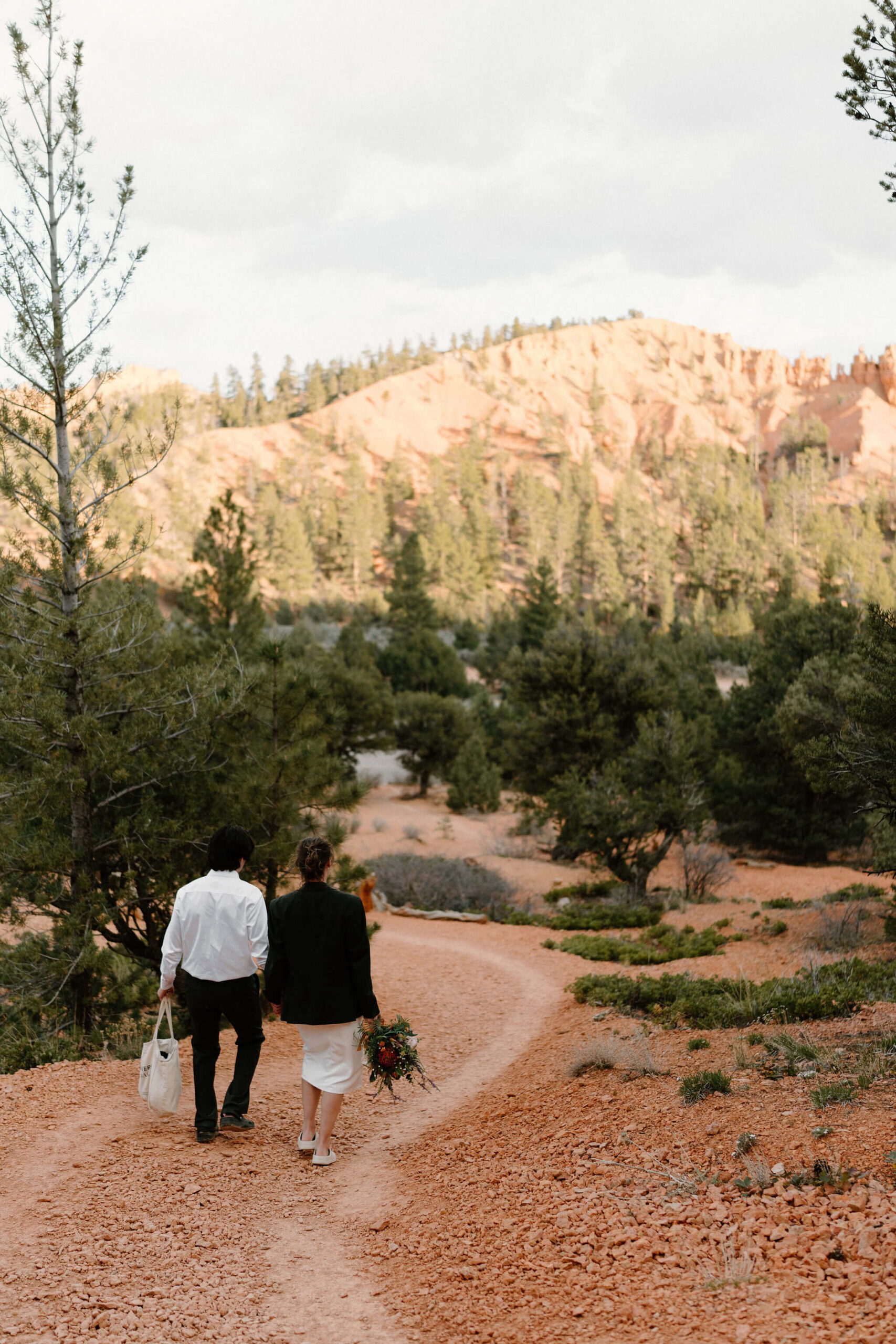 bride and groom walking down a trail, groom holding bride's tote bag and bride wearing groom's jacket and holding her bouquet