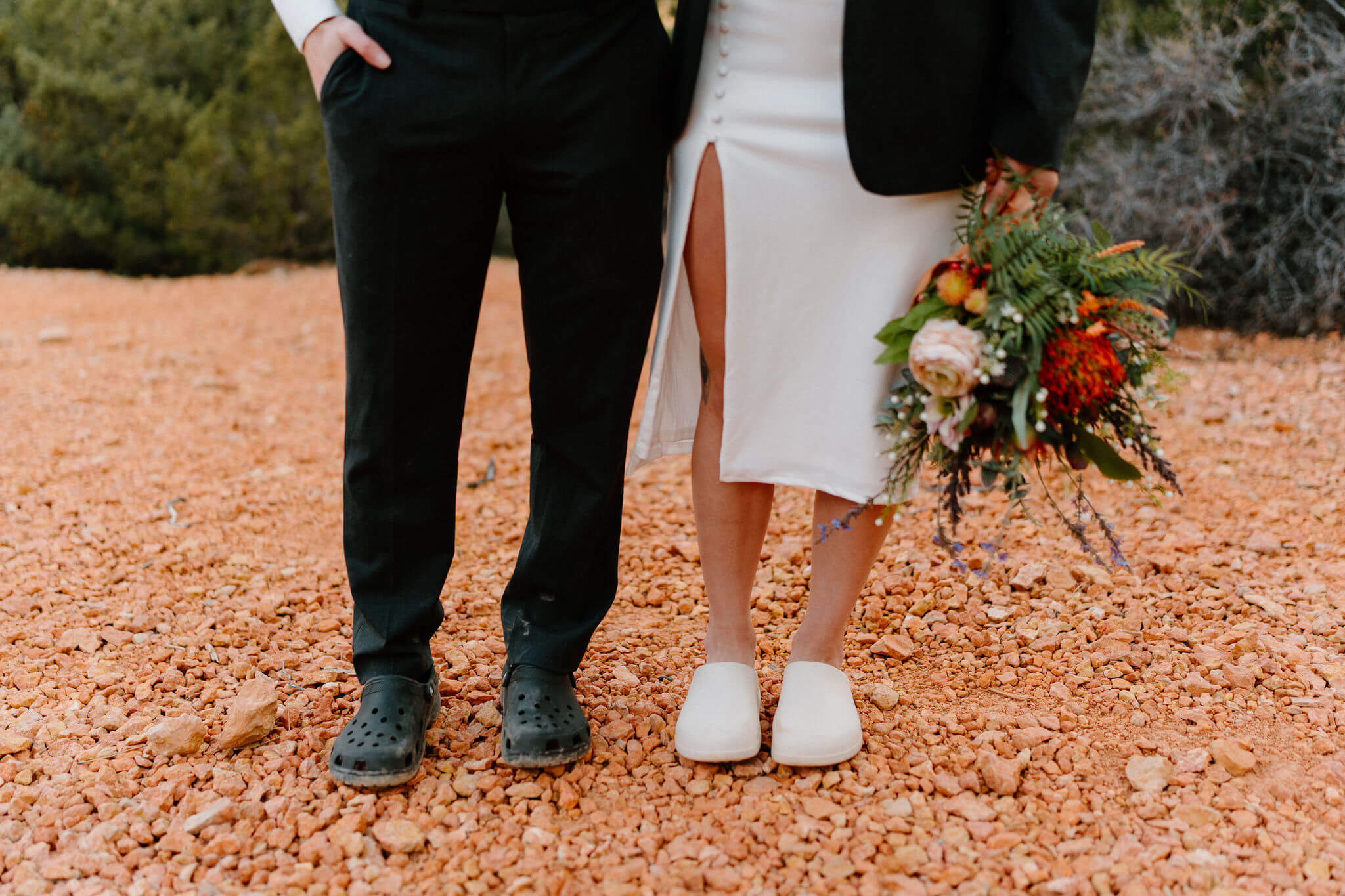 close up of bride and groom from the knee down, both wearing crocs (black and white, respectively), while bride is wearing groom's jacket and holding her bouquet at her side