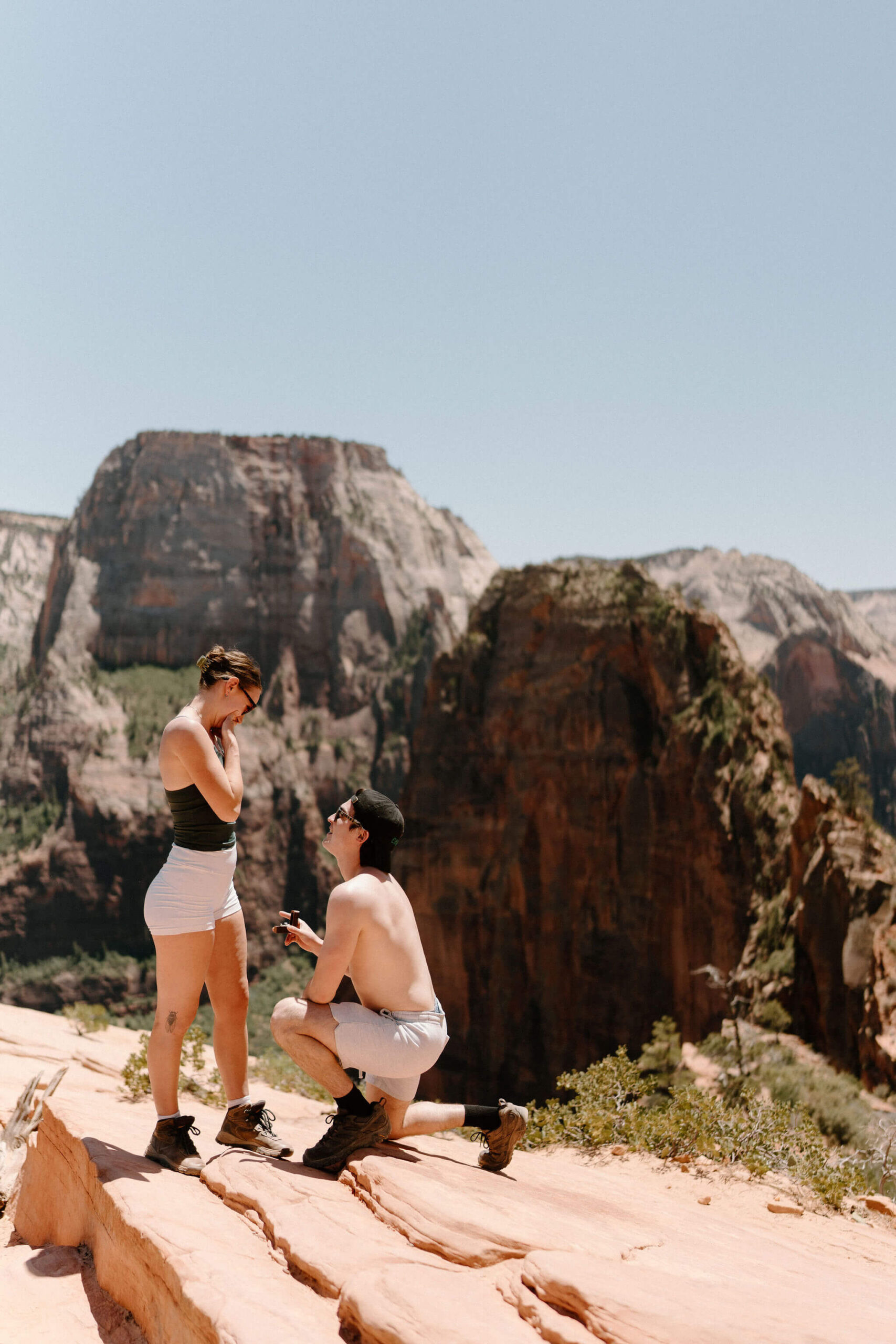 man on one knee proposing to a woman at Scout Lookout, with a view of Angel's Landing hike at Zion National Park on a clear blue day