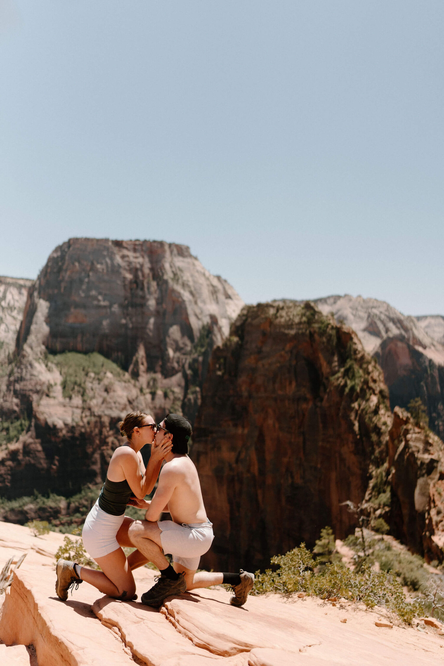 woman leaning down to kiss her new fiance at Scout Lookout, with a view of Angel's Landing hike at Zion National Park on a clear blue day