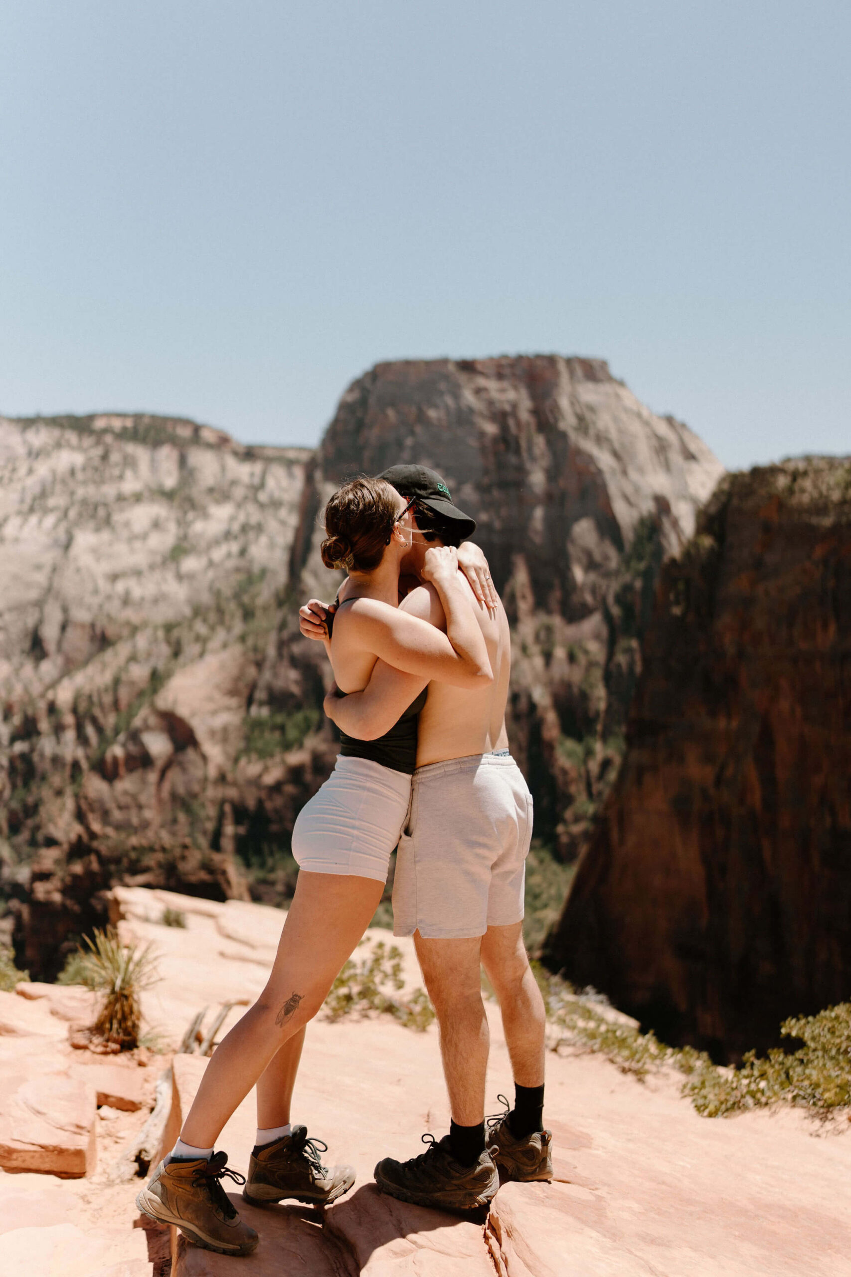 man and woman embracing at Scout Lookout, with a view of Angel's Landing hike at Zion National Park on a clear blue day after just getting engaged on a hike