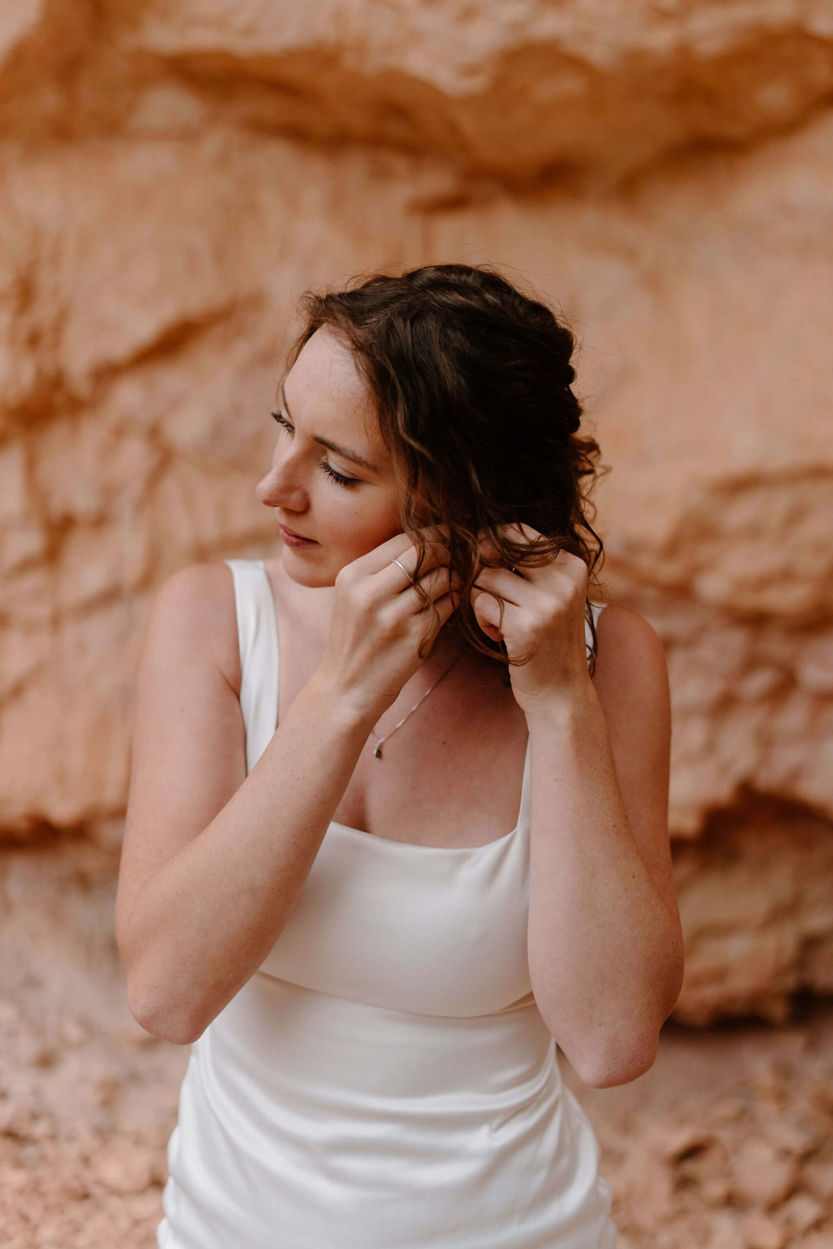 bride in white square neck sleeveless dress putting in earrings against a red rock backdrop