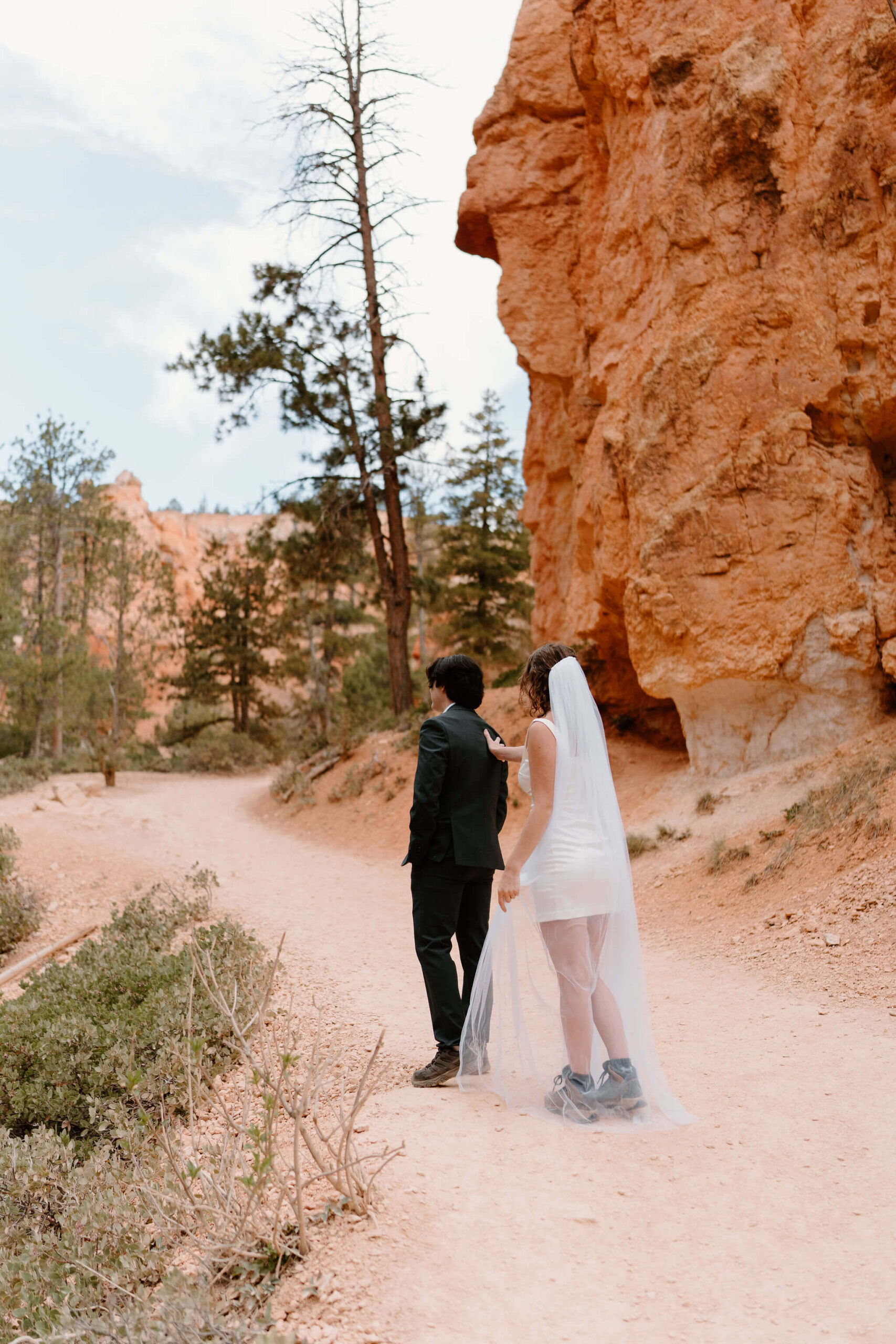 bride in a short white dress, a floor-length veil, and hiking boots walking toward groom (black suit) for their first look on a hiking trail through red rocks and hoodoos