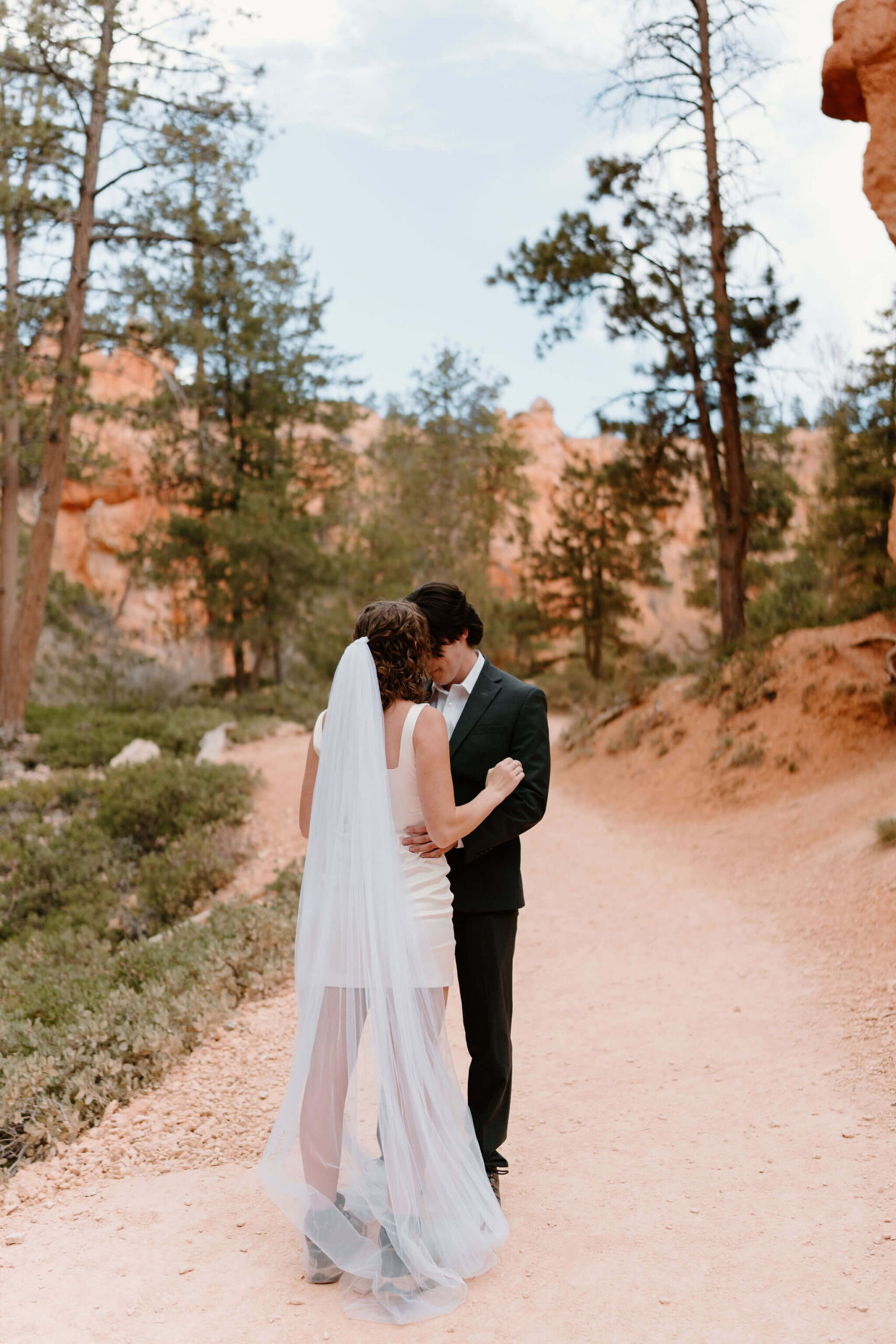 bride (short white dress, floor length veil, hiking boots) and groom (suit, hiking boots) embracing on a hiking trail at Bryce Canyon National Park