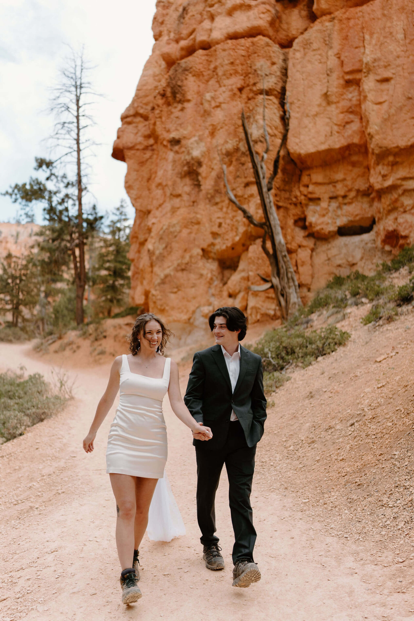 bride (short white dress, floor length veil, hiking boots) and groom (gray suit, hiking boots) holding hands and walking toward the camera down a trail