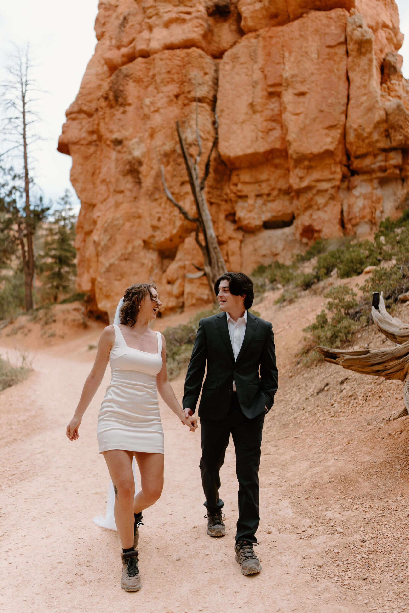 bride (short white dress, floor length veil, hiking boots) and groom (gray suit, hiking boots) holding hands and walking toward the camera down a trail
