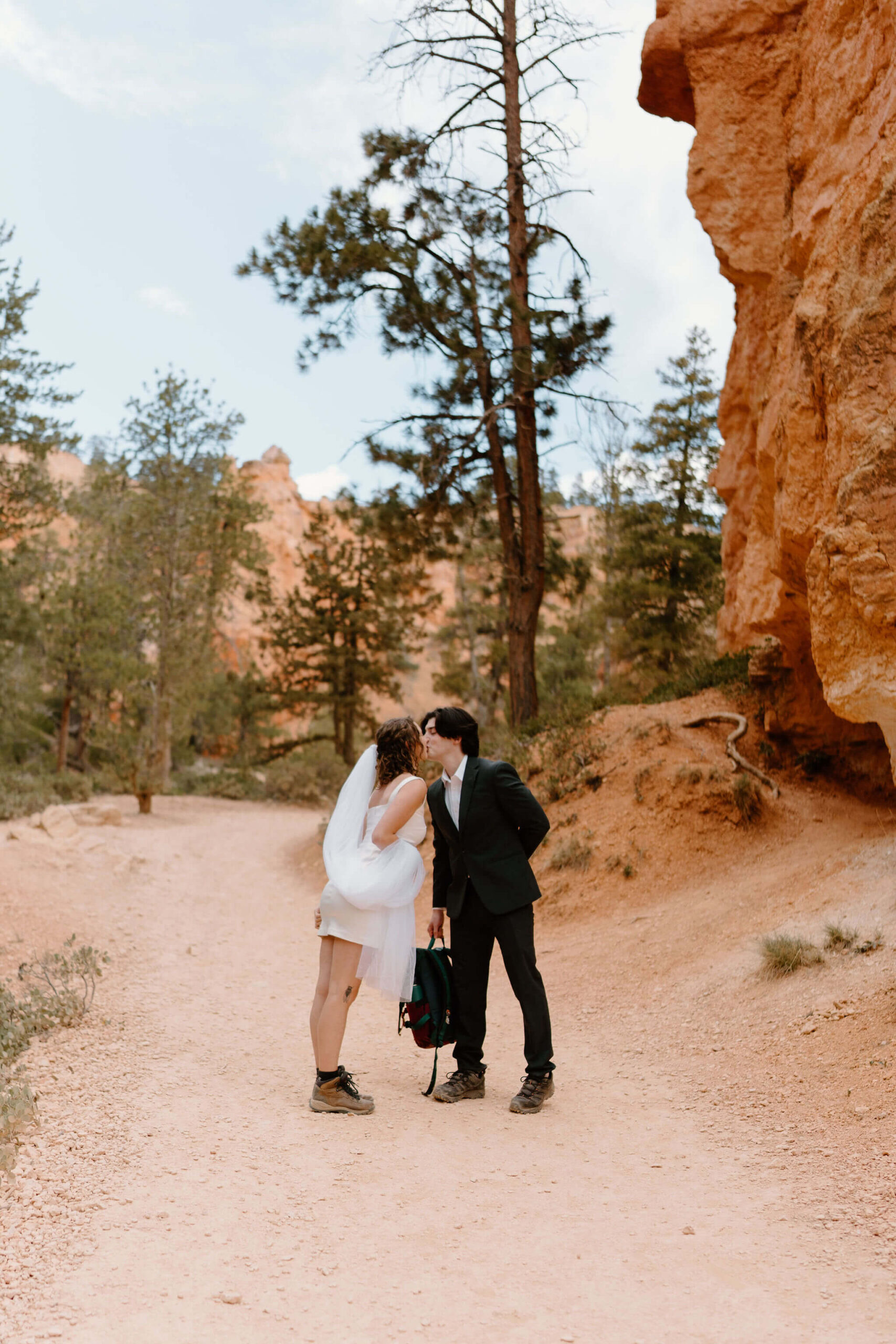 bride and groom wearing hiking boots kissing in the middle of a trail in Bryce Canyon National Park. groom holding their hiking backpack
