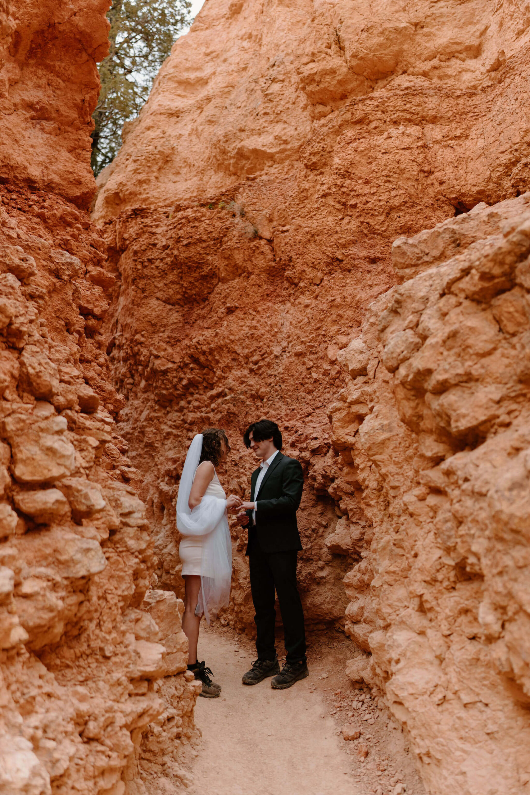 bride and groom on a narrow trail surrounded by tall red rocks, facing each other and exchanging wedding rings