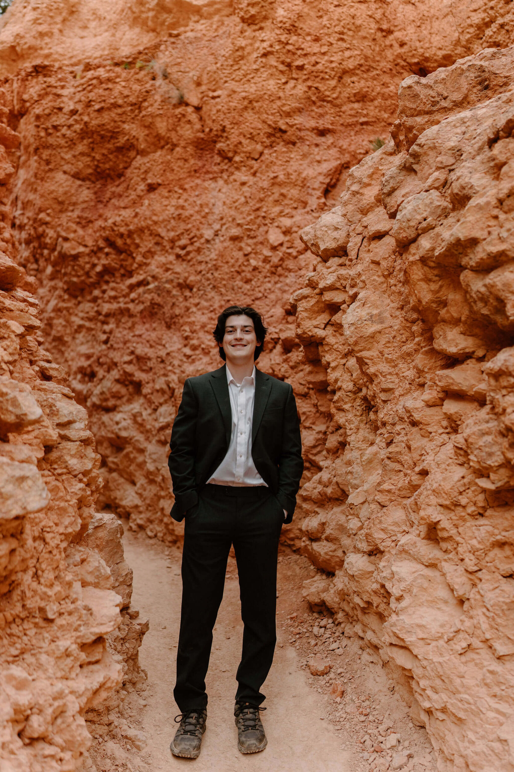 groom on a narrow trail surrounded by tall red rocks, facing the camera and smiling with his hands in his pockets