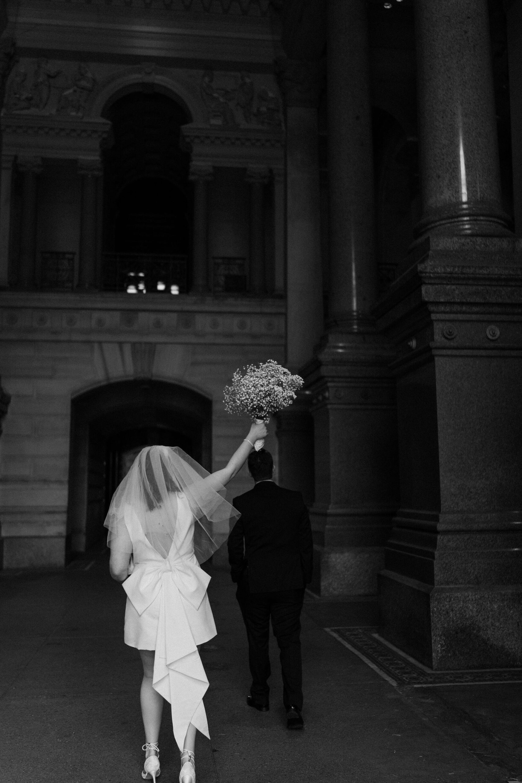 black and white image of a bride (short white dress with a bow) holding up her baby's breath bouquet, next to groom, walking away from the camera through a large building
