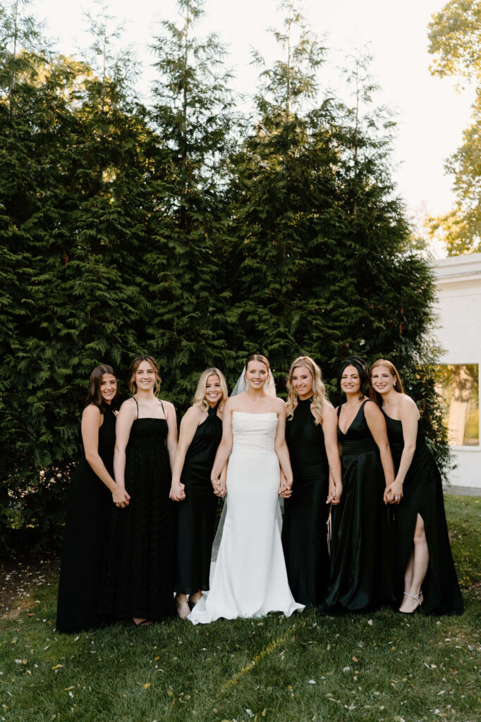 bride (white strapless wedding gown) and bridesmaids (variety of black dresses) holding hands and smiling at the camera