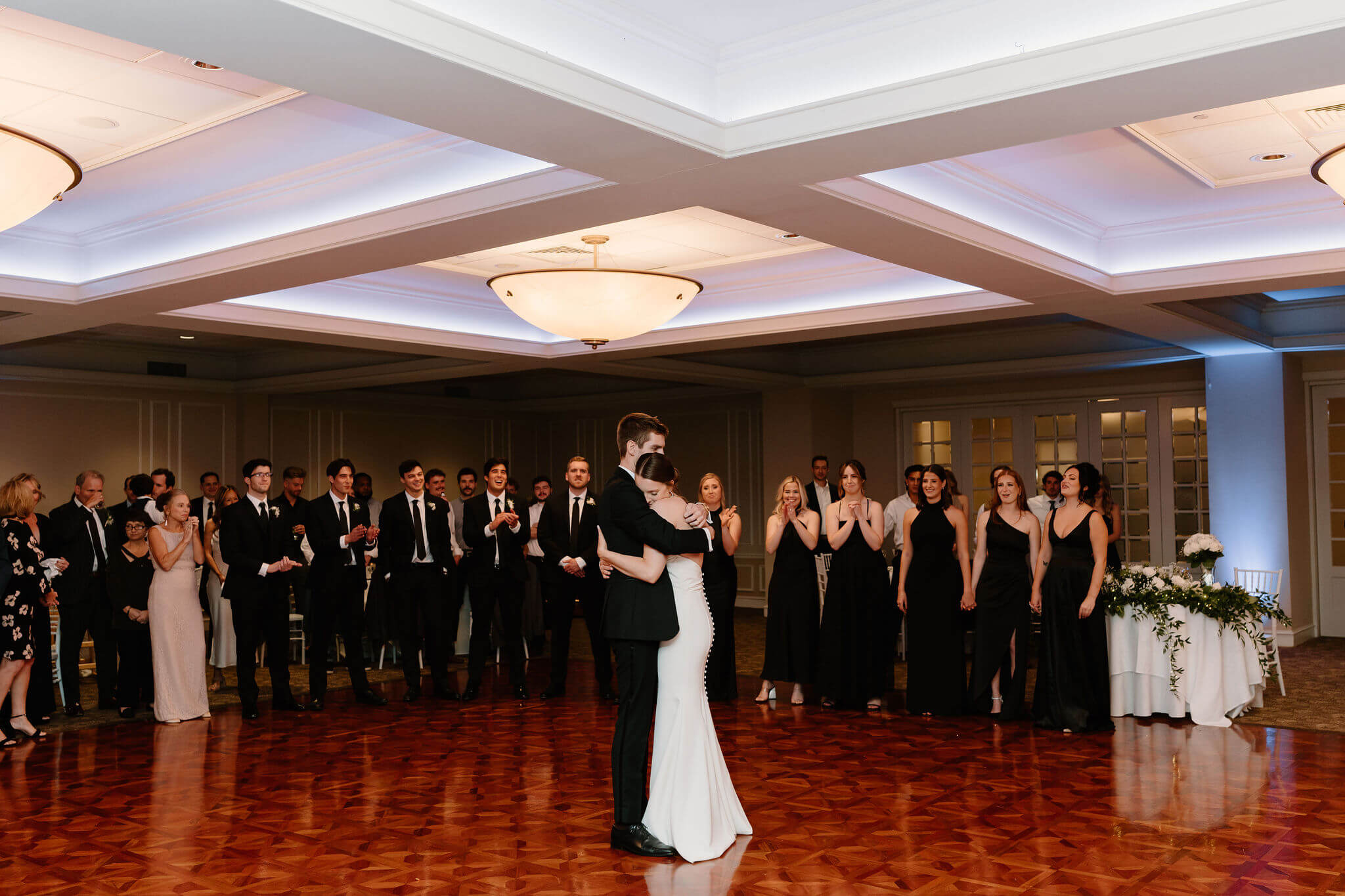 bride and groom embracing closely during their first dance, with their wedding party watching behind them.