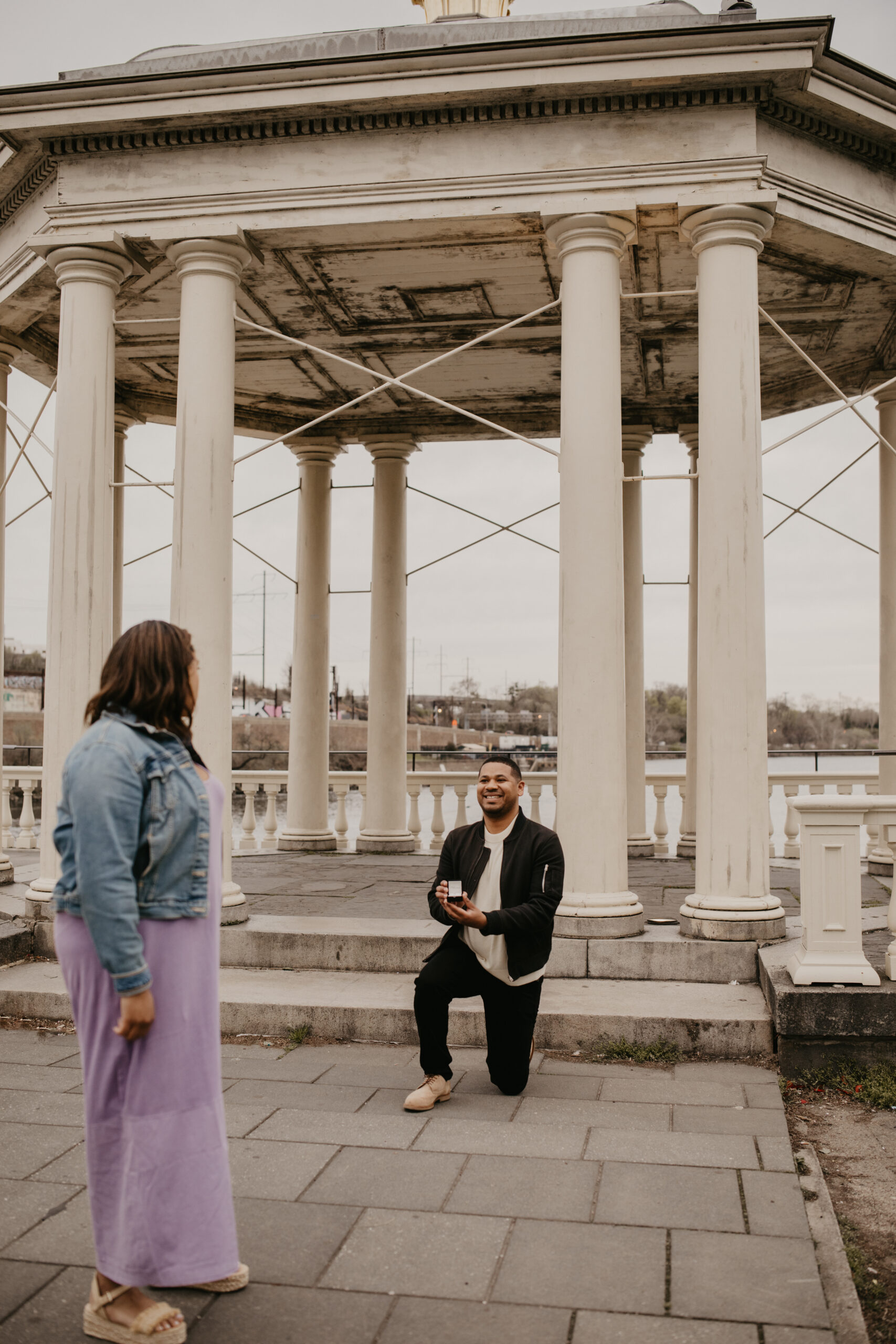 man on one knee proposing to his girlfriend in front of a white waterfront pavilion, she is just turning around to see