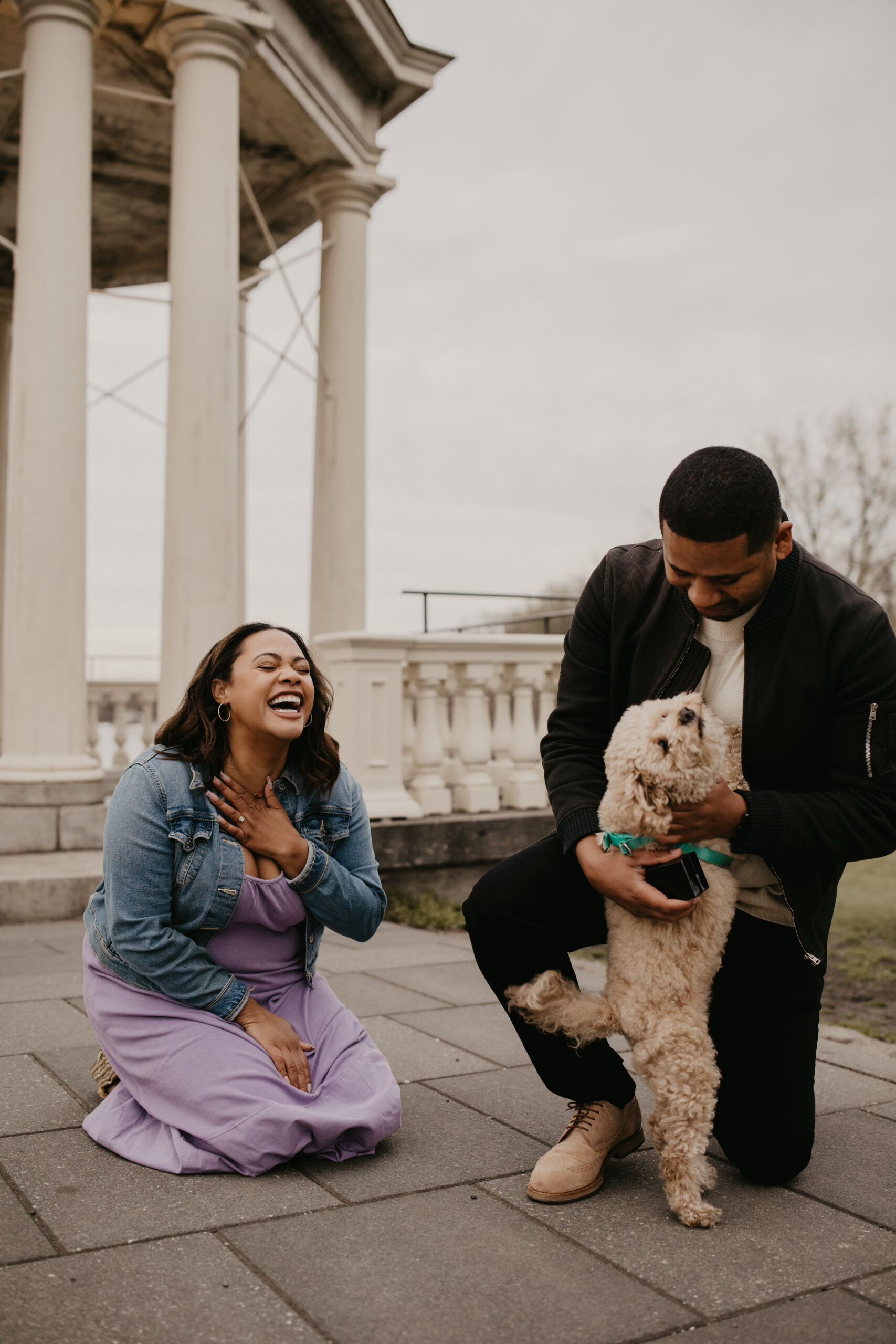 man kneeling down petting his dog, while his new fiancee is kneeling holding her left hand to her chest and laughing with joy