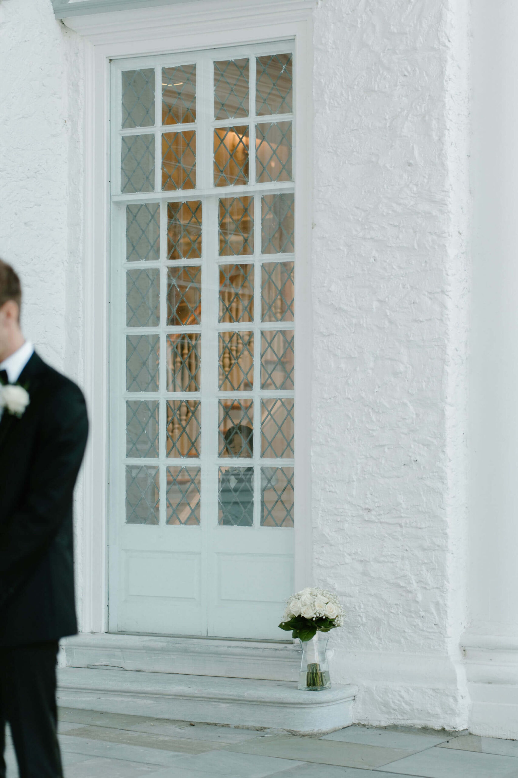 a young boy looking out of a window, photographed from outside, watching the bride and groom exchange wedding vows. The bride's bouquet is on a ledge in front of the window.