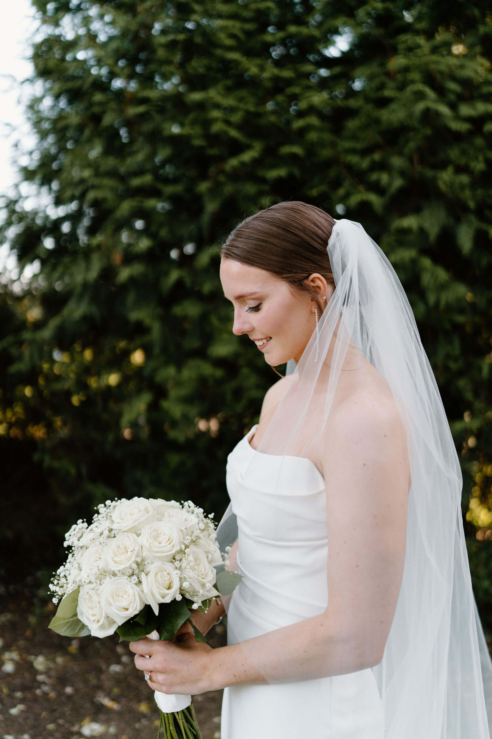 bride with a long veil in a white strapless wedding dress, smiling softly down at her white rose bouquet
