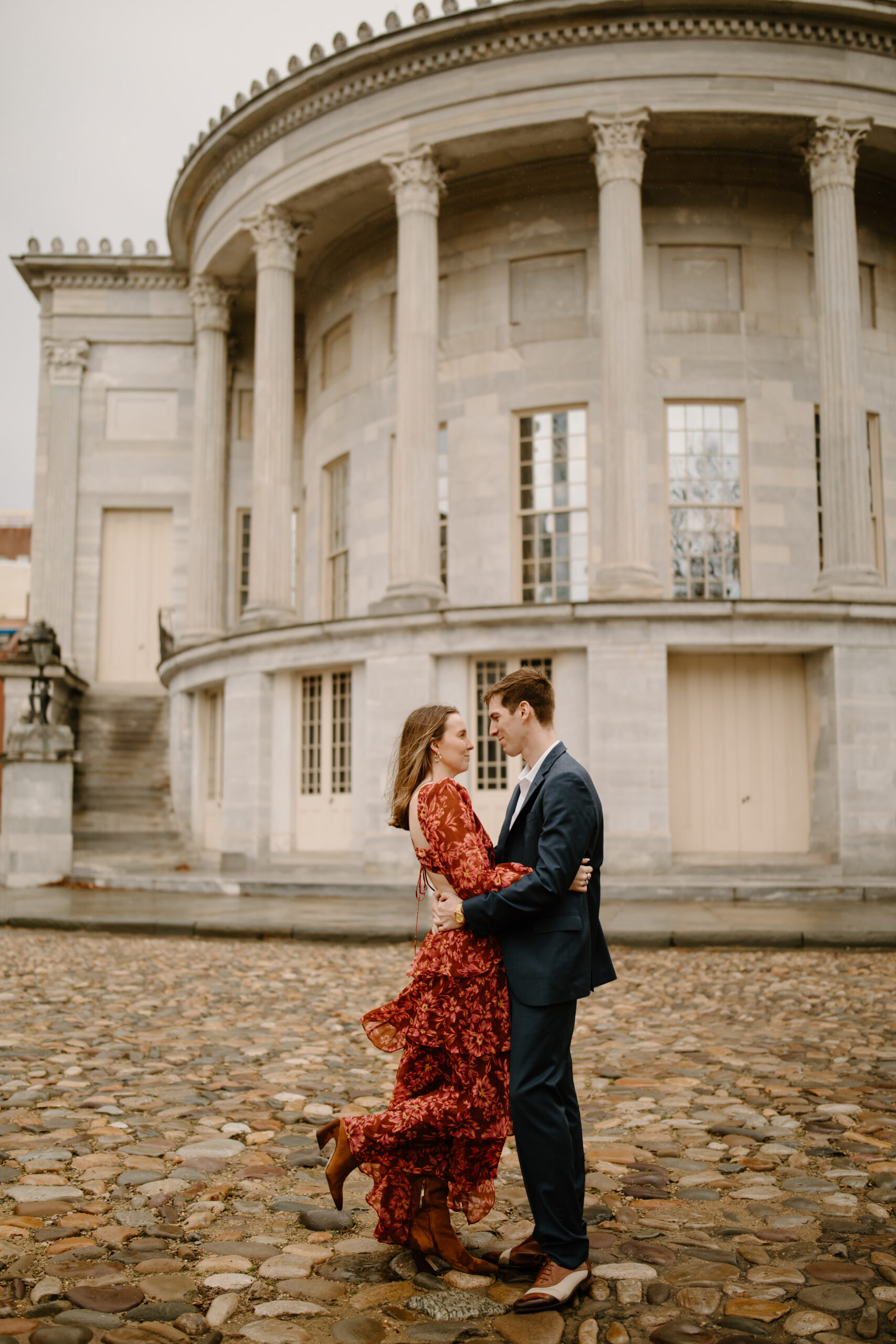 Couple embracing, her heel kicked up in joy, in front of the Merchant's Exchange Building in Philadelphia for engagement photos