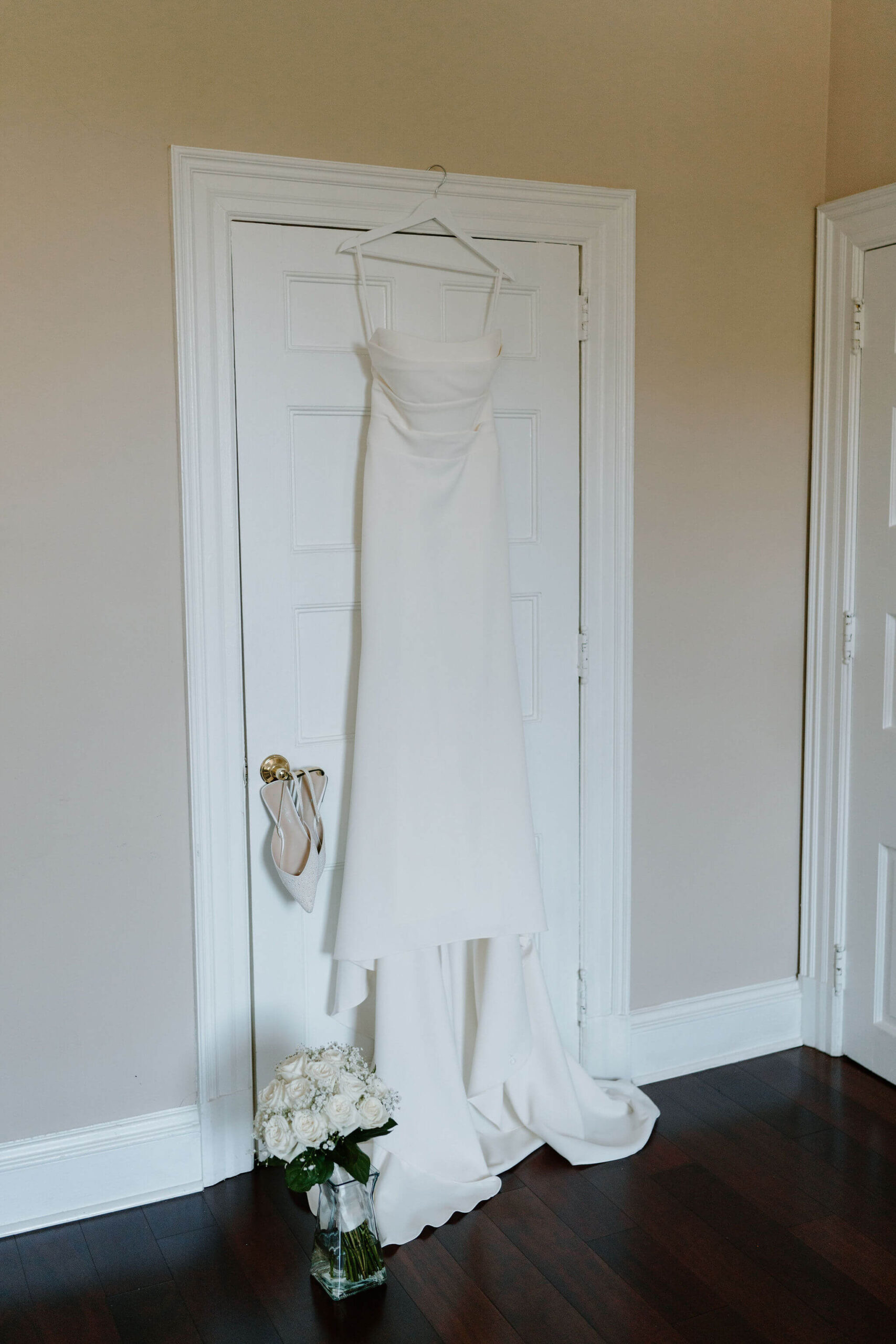 wedding gown hanging from a door frame, shoes hanging from the handle, and a white bouquet in a vase on the floor of a bridal suite