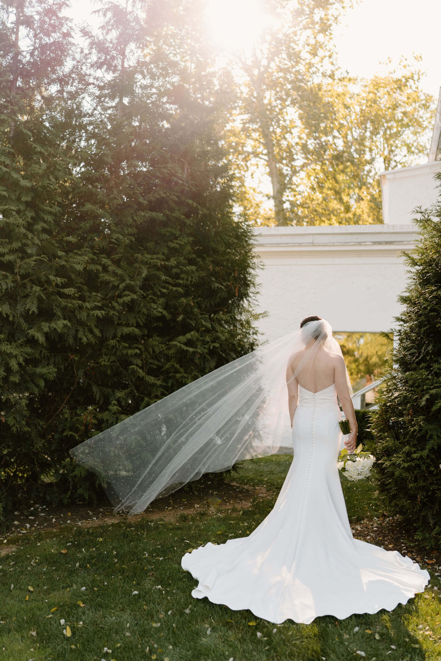 bride in a long white backless wedding dress and a long veil blowing in the wind, facing away from the camera with her bouquet held down at her side
