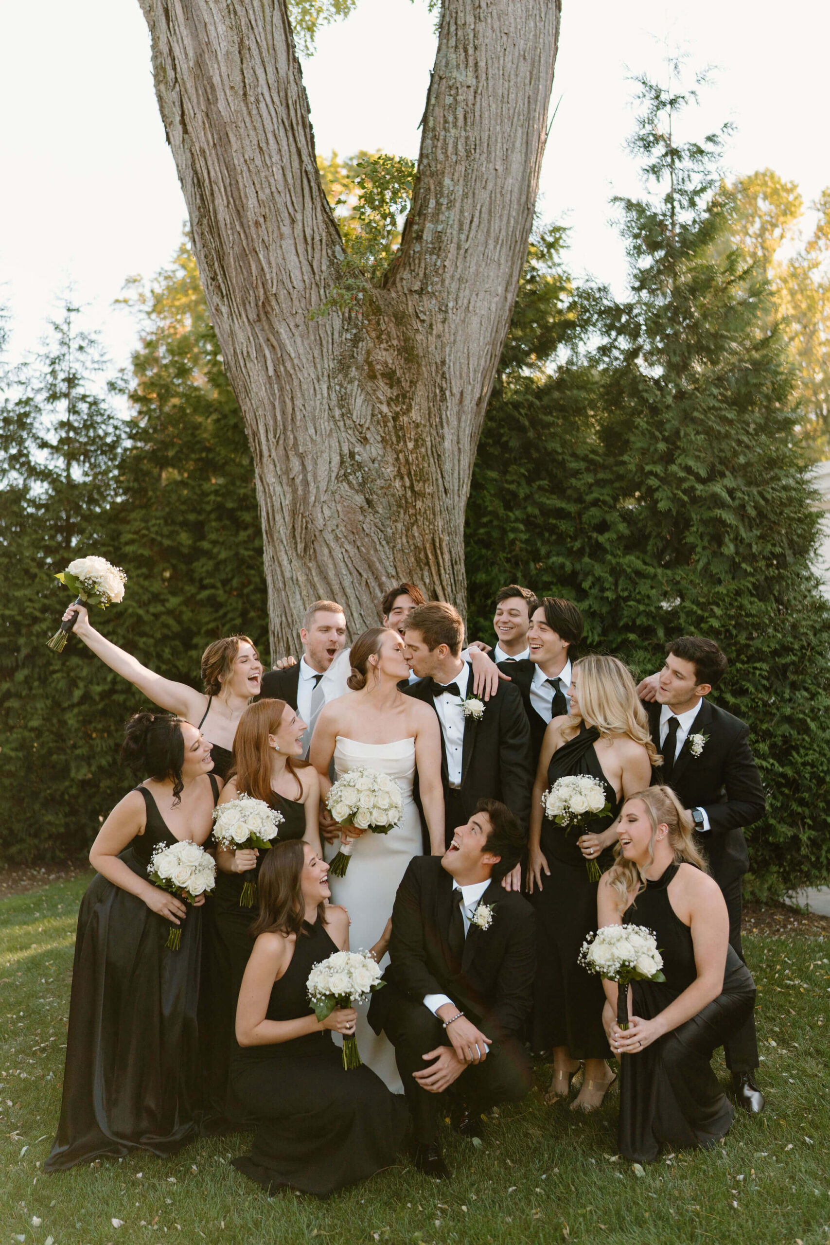 bride and groom (white wedding dress, black suit, respectively) kissing while surrounded by their wedding party (all wearing black dresses and suits with white roses), cheering toward the couple