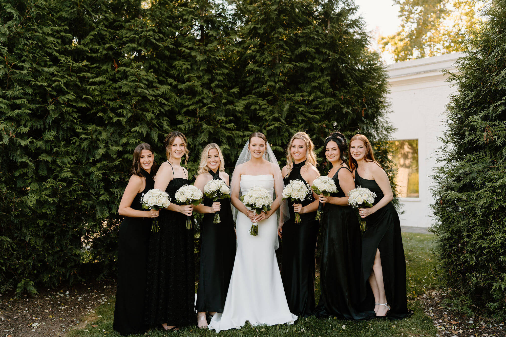 bride (long white gown, long veil) and bridesmaids (all black dresses in a variety of styles), standing next to each other and smiling at the camera while holding bouquets of white roses