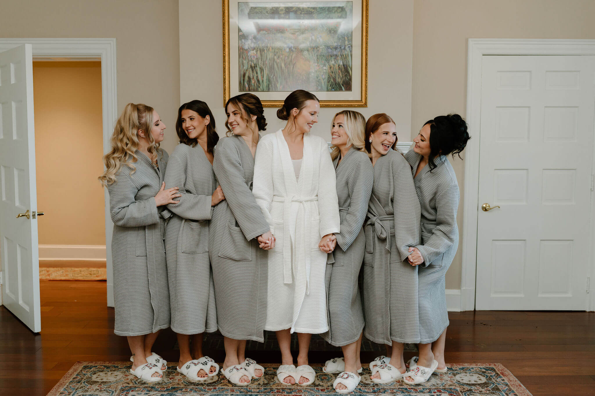 bride (white robe) with six bridesmaids (gray robes), holding hands and smiling at each other wearing matching white slippers