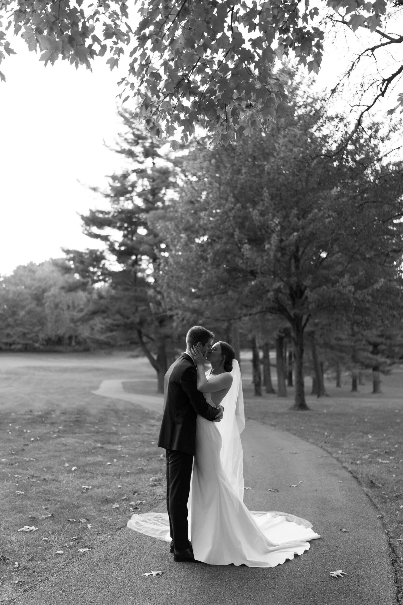 Black and white image of bride and groom embracing and kissing on a path through trees