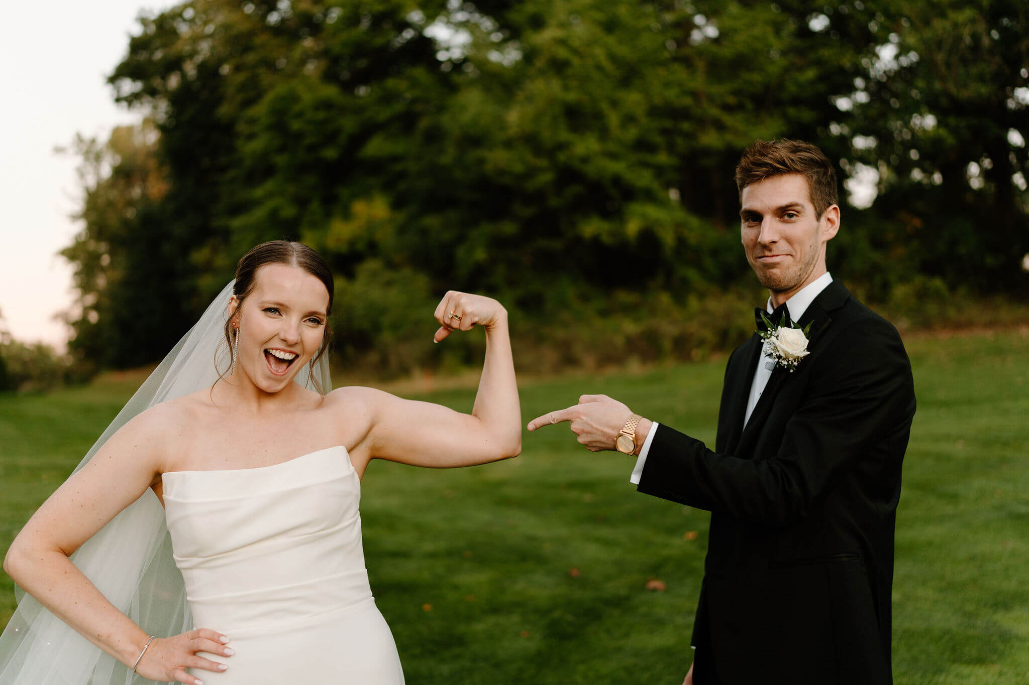 groom pointing at bride's flexed bicep muscles, while she smiles proudly at the camera