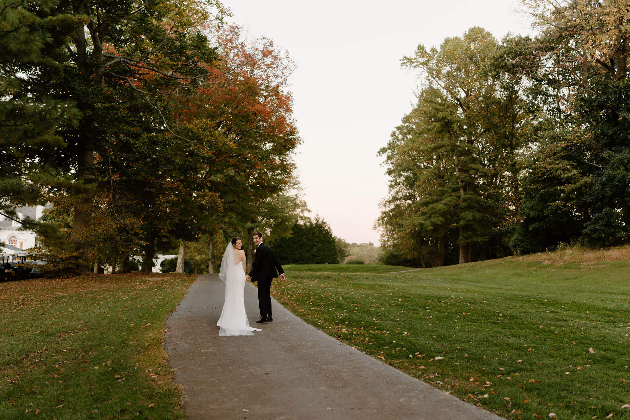 bride and groom holding hands and walking away down a path to a tree with autumn foliage, looking back at the camera and smiling