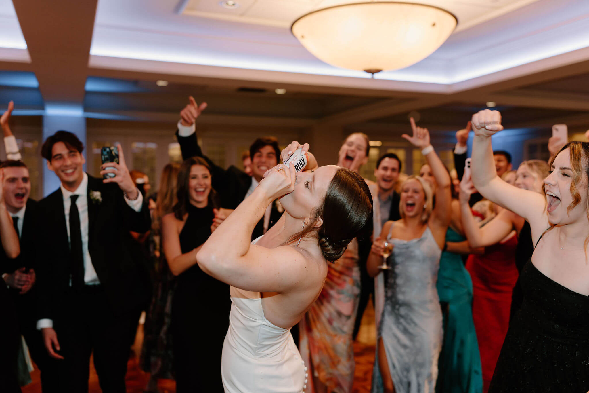 bride shotgunning a canned drink on the dance floor at her wedding reception, surrounded by guests cheering and waving