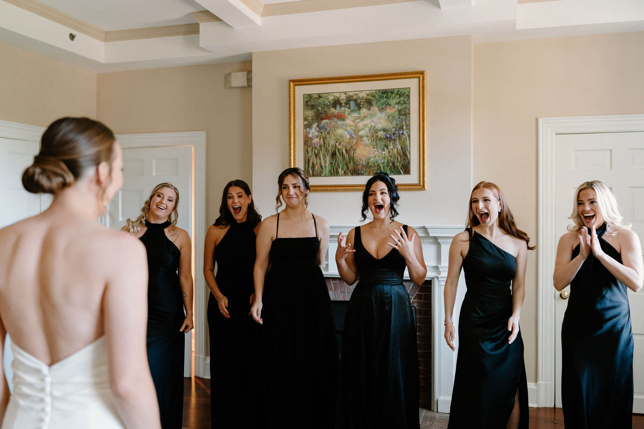 first look of a bride with her bridesmaids, all wearing black dresses, smiling with shock toward the bride (facing away from camera)