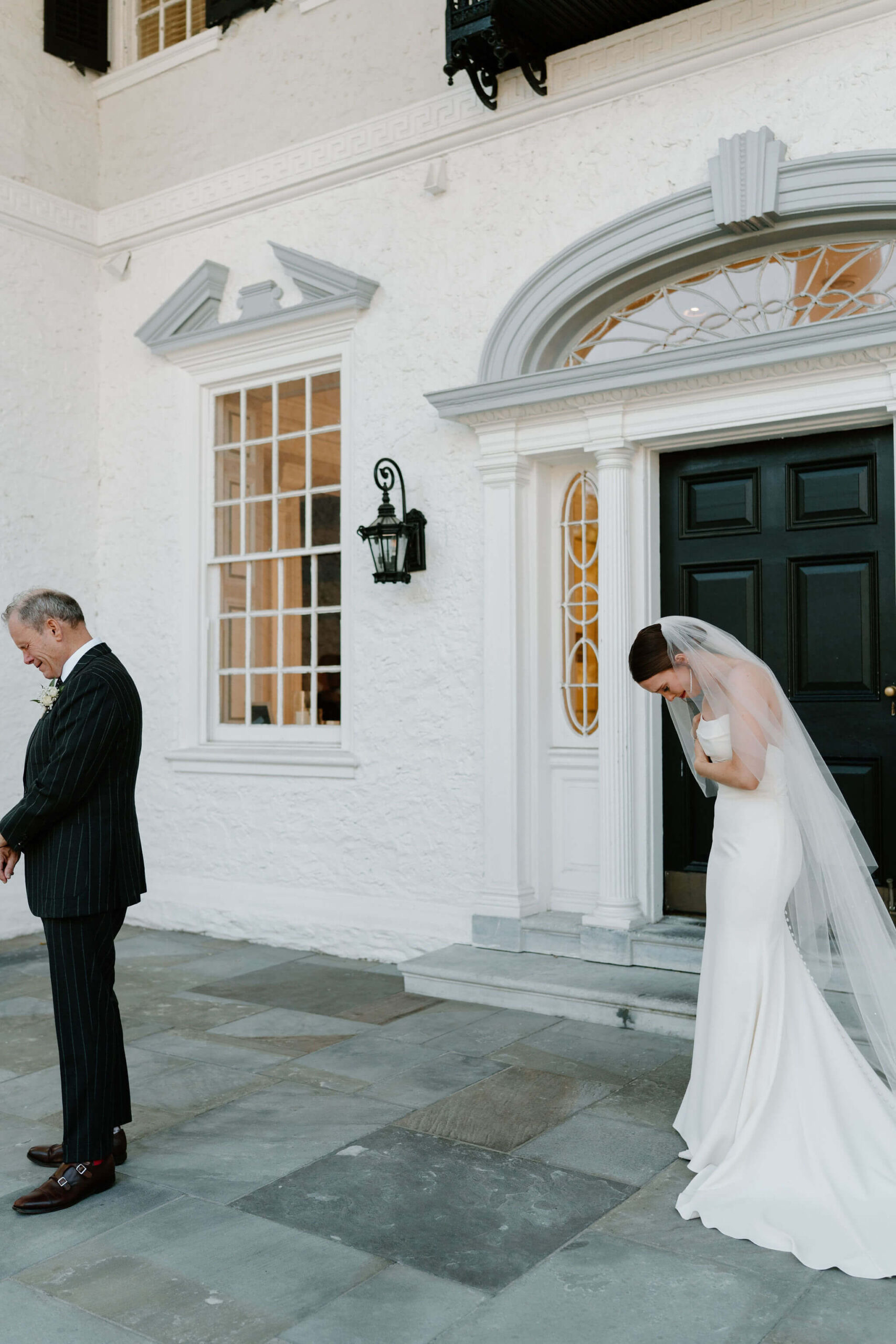 bride in a white wedding dress with a train and a floor length veil hunched over and crying before her first look with her father, standing with his back to her in a suit.