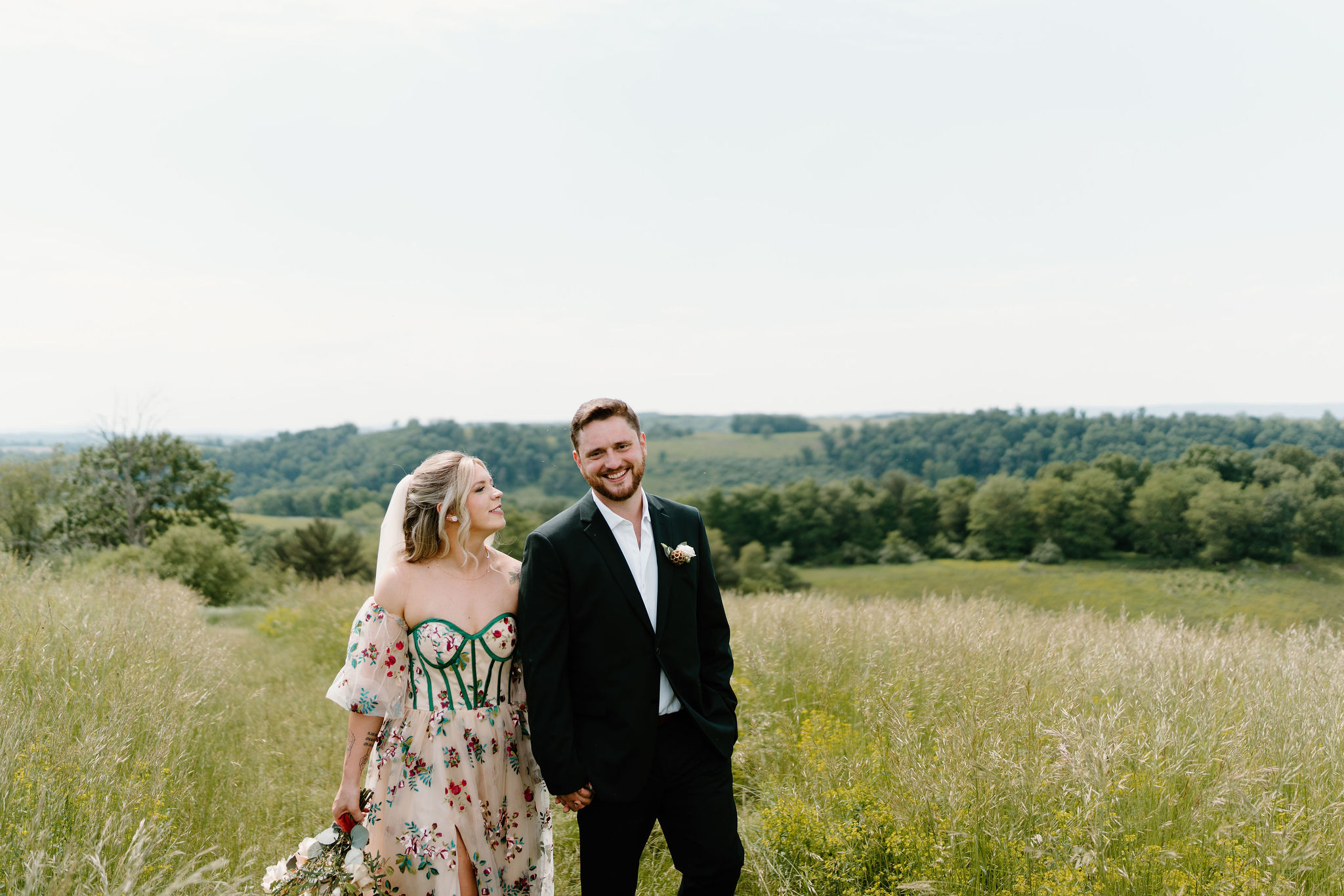 bride wearing a nontraditional wedding dress smiling at her groom, who looks at the camera, as they walk through a tall grassy field on their wedding day
