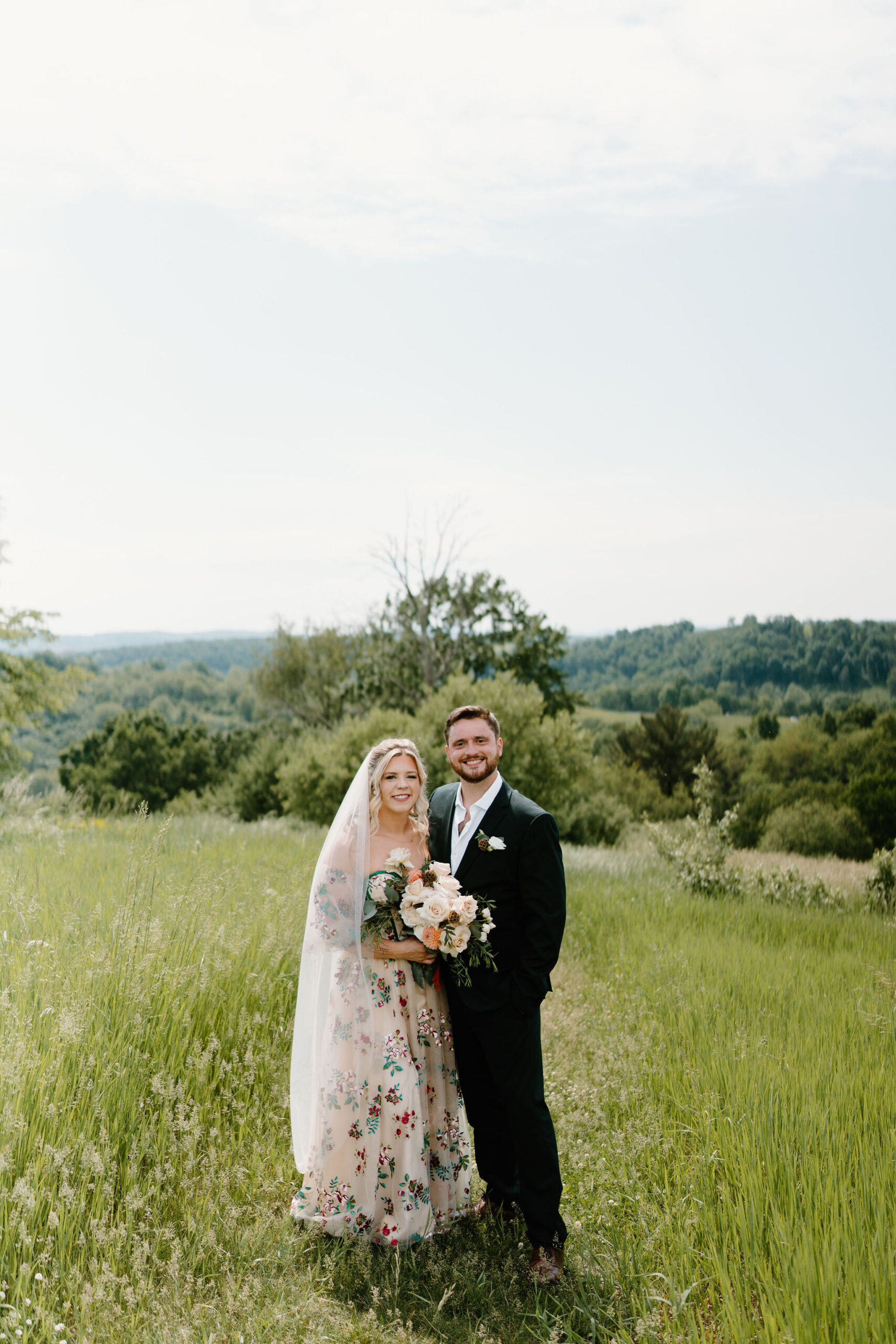 bride in a colorfully embroidered wedding gown and a long veil, holding a bridal bouquet and standing next to her groom (dark green suit), smiling at the camera in a tall green field
