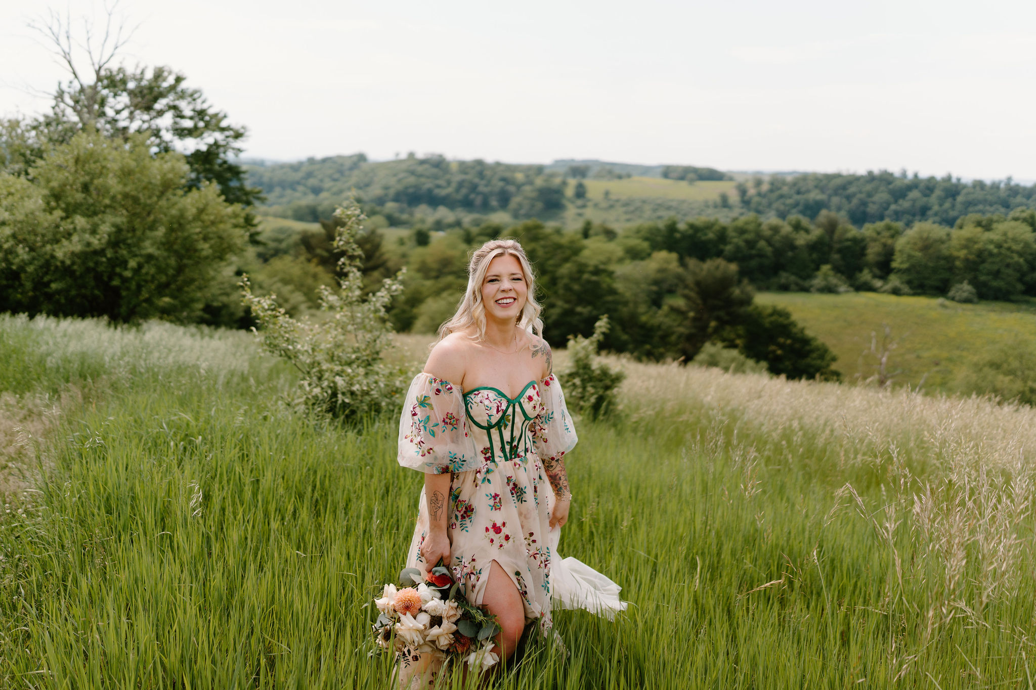 bride with tattoos and an alternative wedding dress smiling at the camera in a tall green field