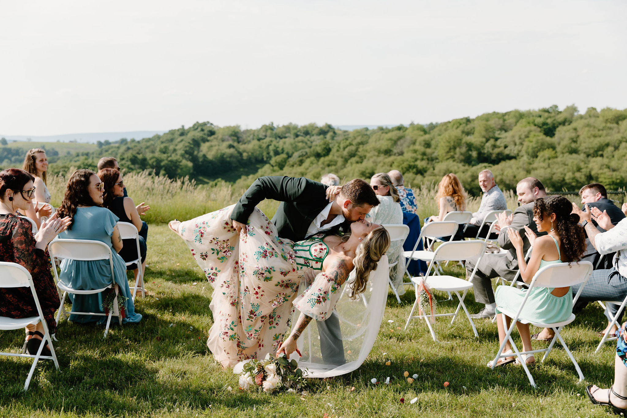 groom dipping his bride in the middle of the aisle after their wedding ceremony, her colorful dress lit up by the sun and bouquet down at her side