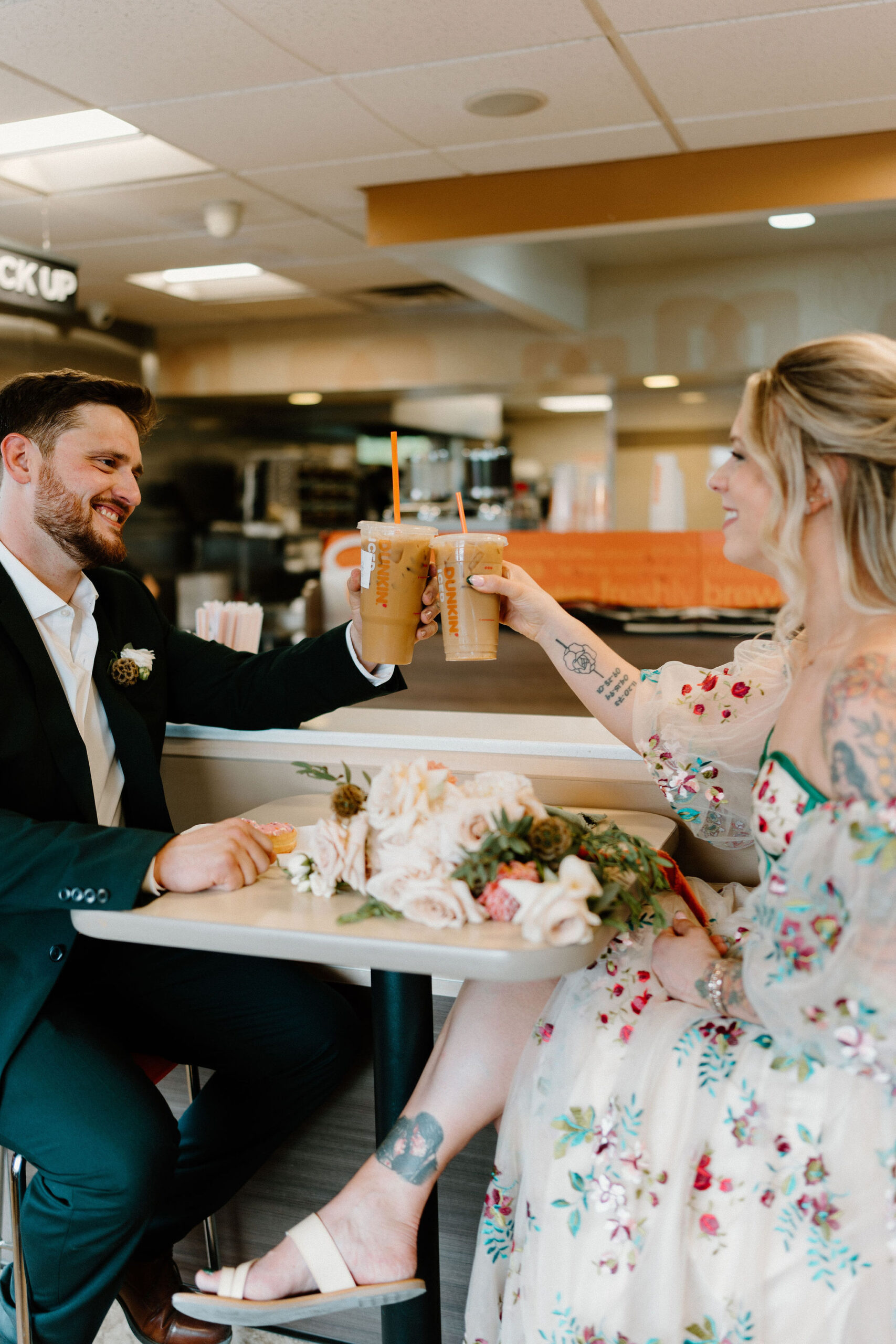 bride and groom tapping their iced coffees in a "cheers", sitting across from each other and smiling in a Dunkin Donuts