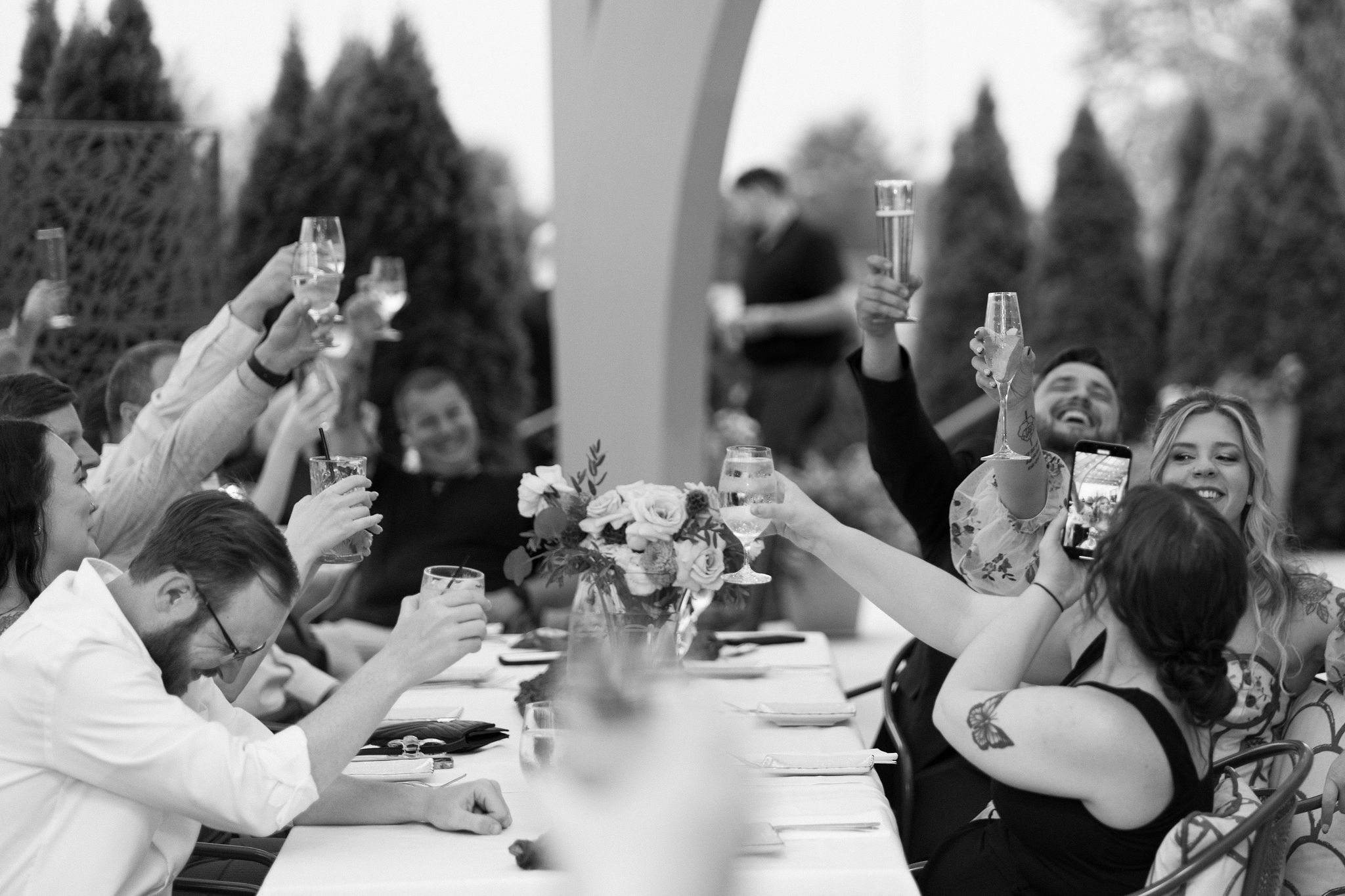 black and white image of bride, groom, and their table laughing and toasting after a speech during the reception