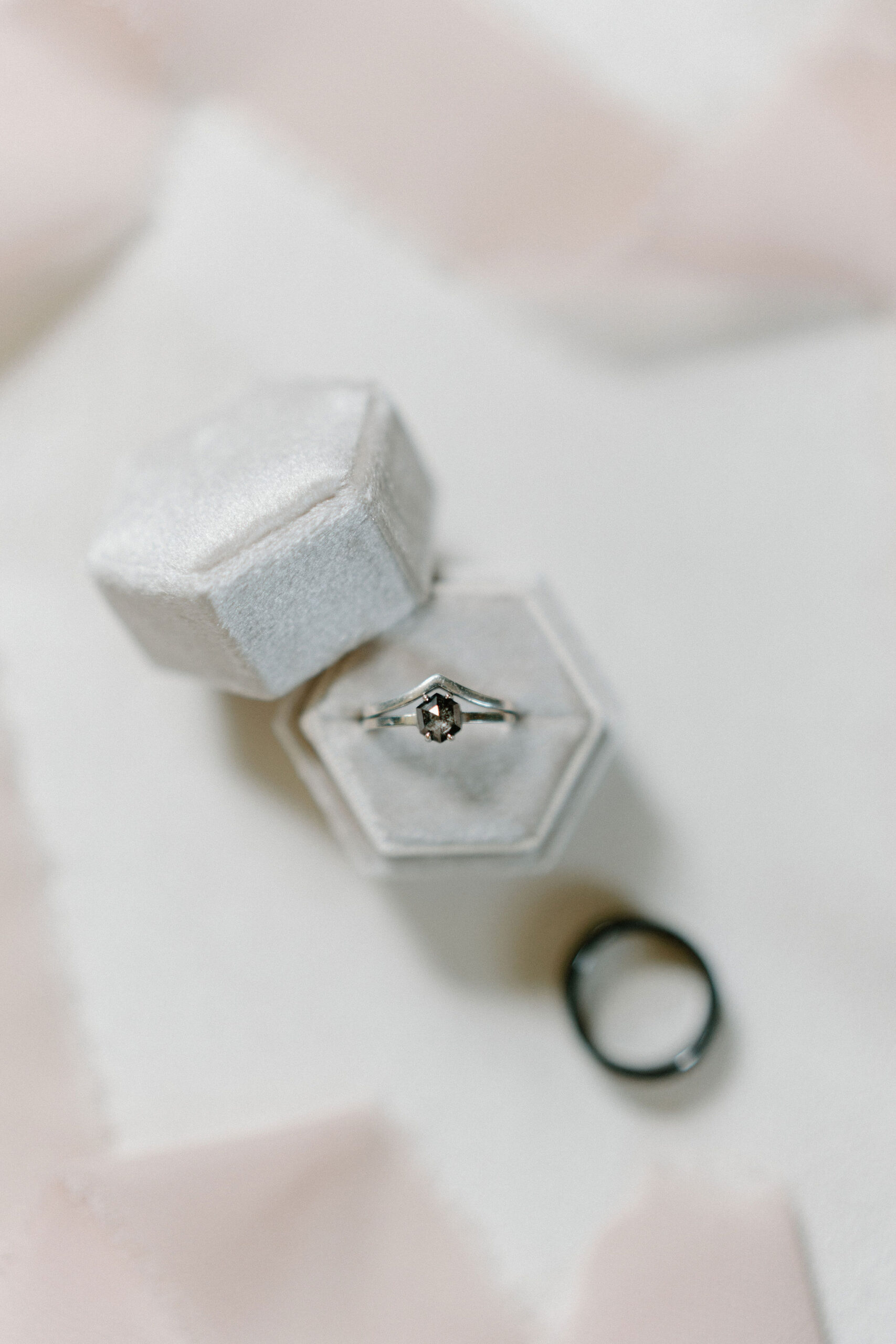 wedding details image of a gray stoned silver engagement ring, a silver wedding band, and a black wedding band, arranged in a white ring box